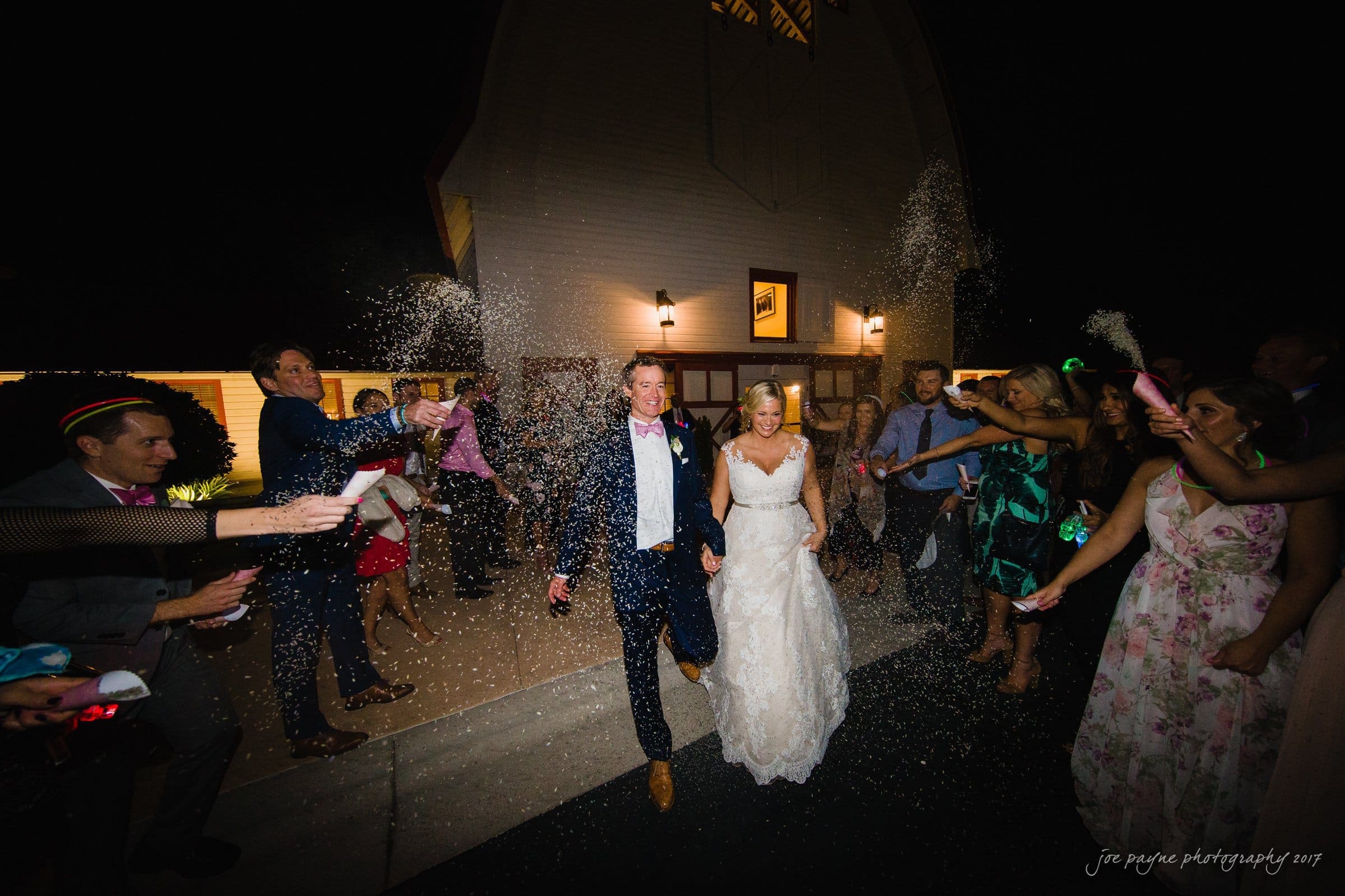 winmock nc wedding photographer - hallie & cullen A newlywed couple in wedding attire joyfully walks through a crowd of guests throwing confetti outside a barn at night. The guests are smiling and celebrating.