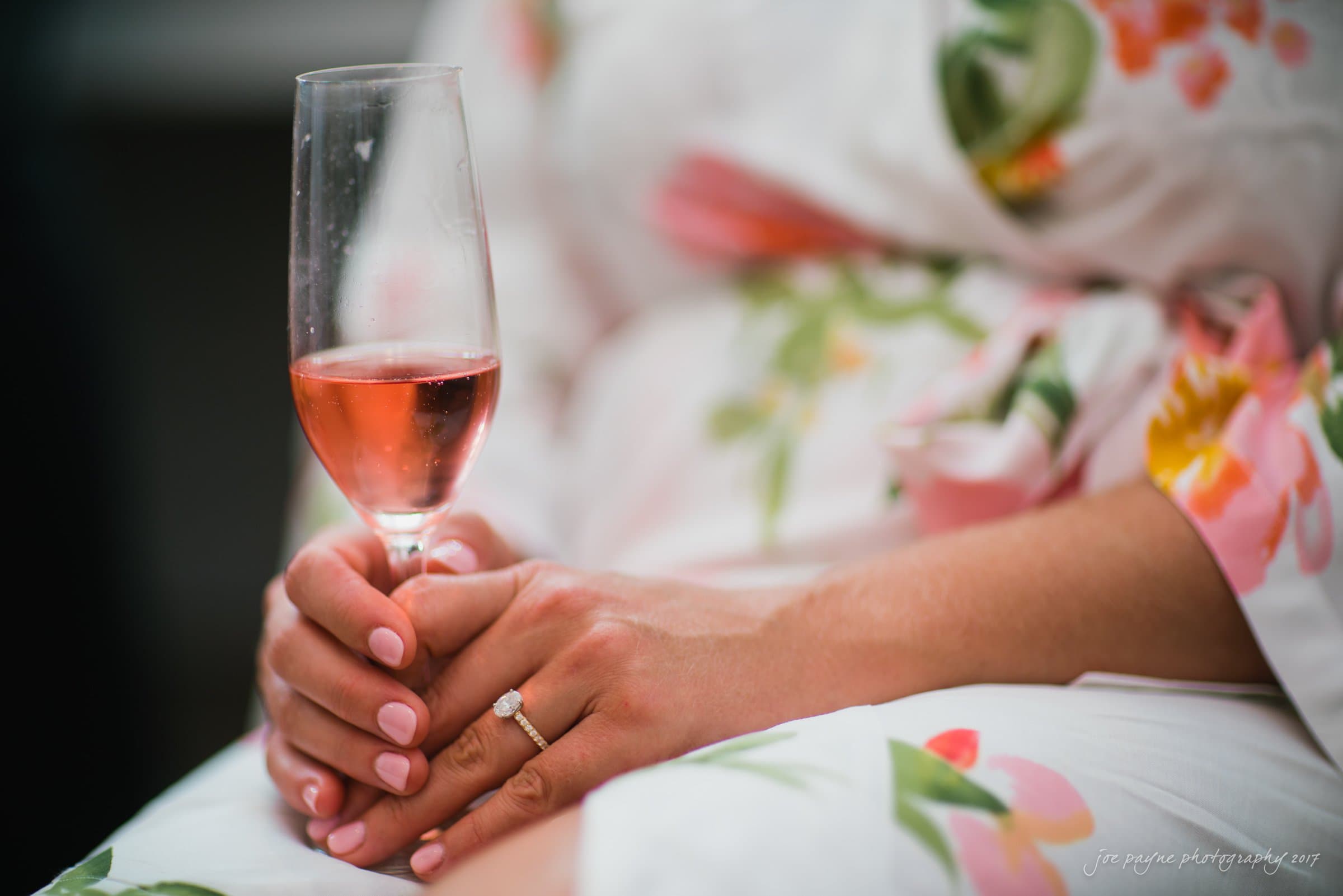 winmock nc wedding photographer - hallie & cullen A person with manicured nails holds a half-filled glass of pink wine. They are wearing a floral-patterned robe, and a ring is visible on their finger. The focus is on the hands and glass, with a soft, blurred background.
