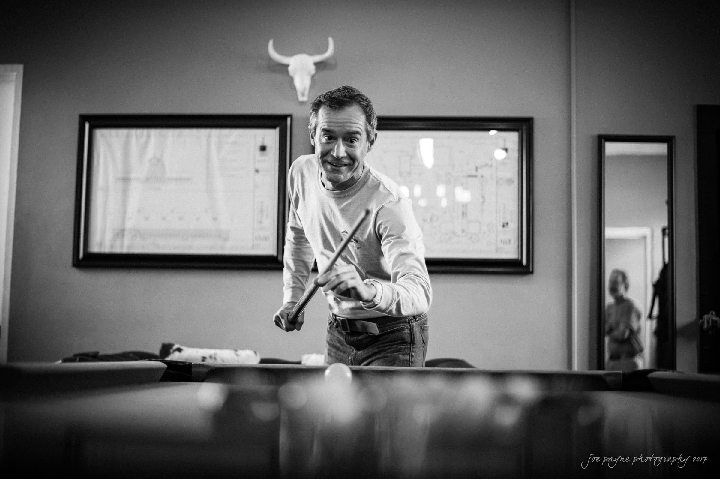 winmock nc wedding photographer - hallie & cullen A man in a casual sweater aiming a cue stick towards the viewer in a billiards room. The room is decorated with a bull skull on the wall and framed documents. The black and white photo captures a friendly moment.