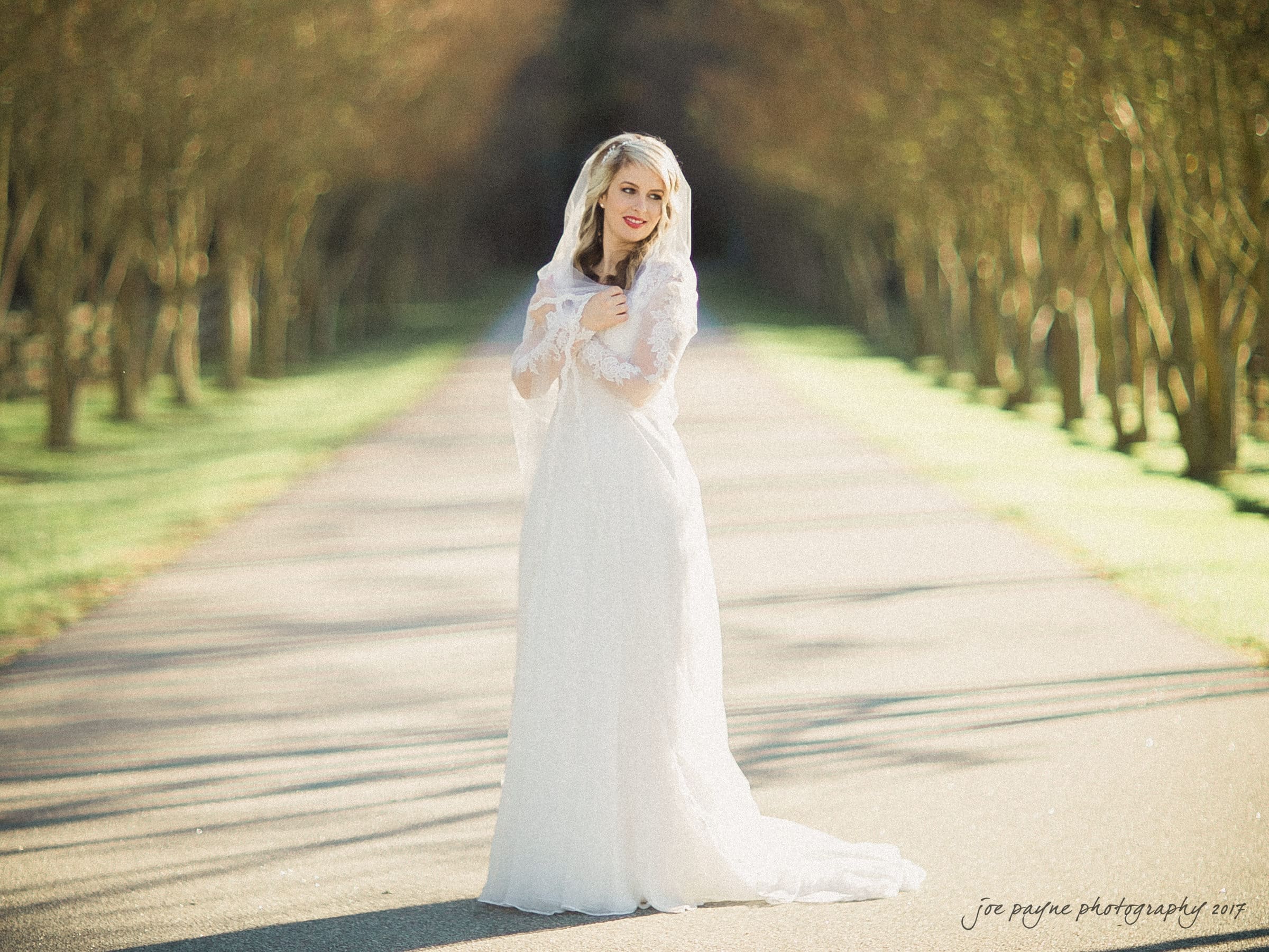 A bride in a white wedding dress stands on a sunlit path lined with trees, smiling and holding her veil. Captured by a Durham wedding photographer, the trees create a natural frame around her, and the soft lighting adds a dreamy atmosphere to the scene.