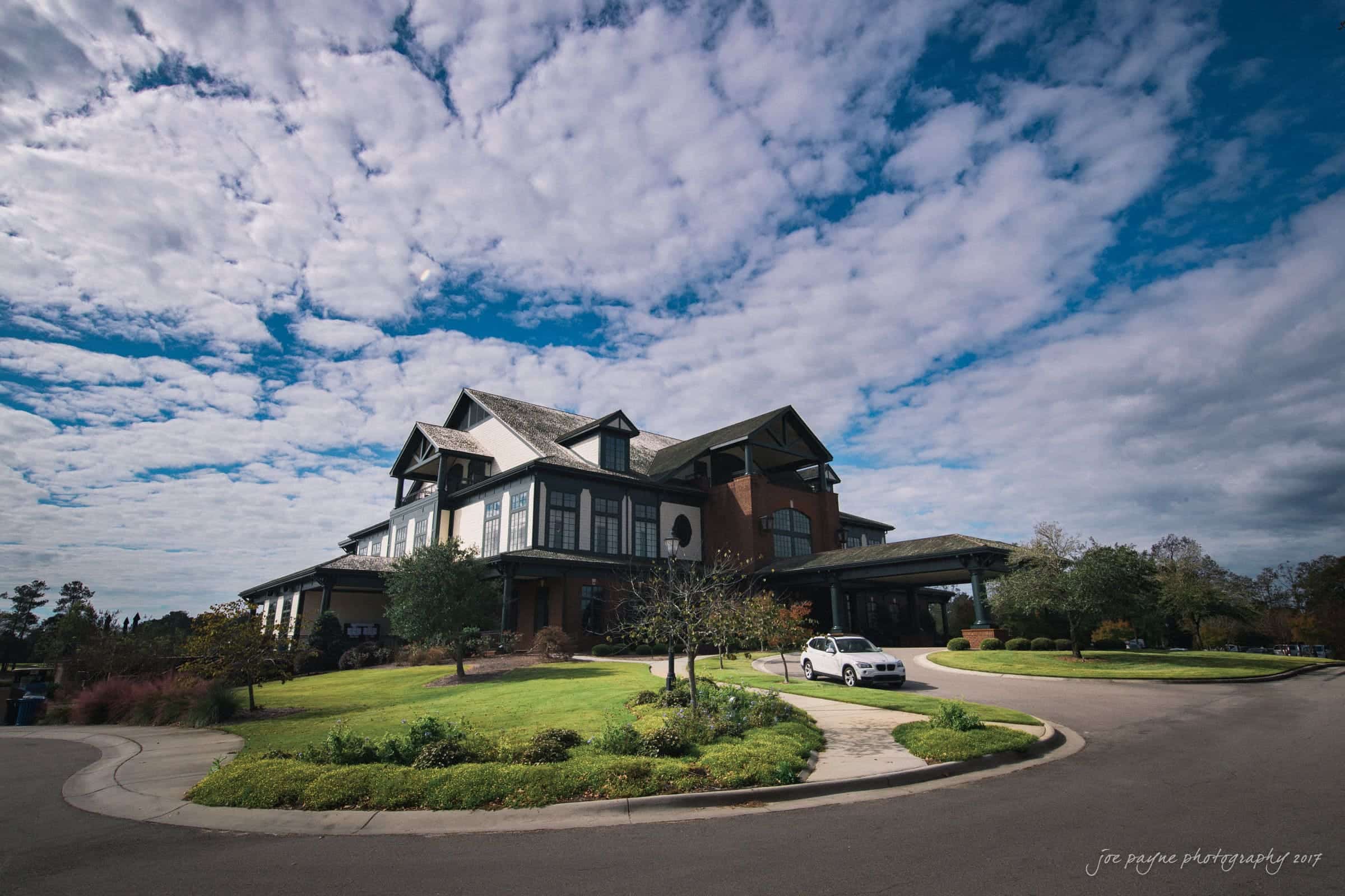 river landing country club wedding - megan & chad Large two-story house with a dark roof and brick accents, surrounded by green lawns and trees. A white car is parked in the driveway. The sky above is partly cloudy with patches of blue.