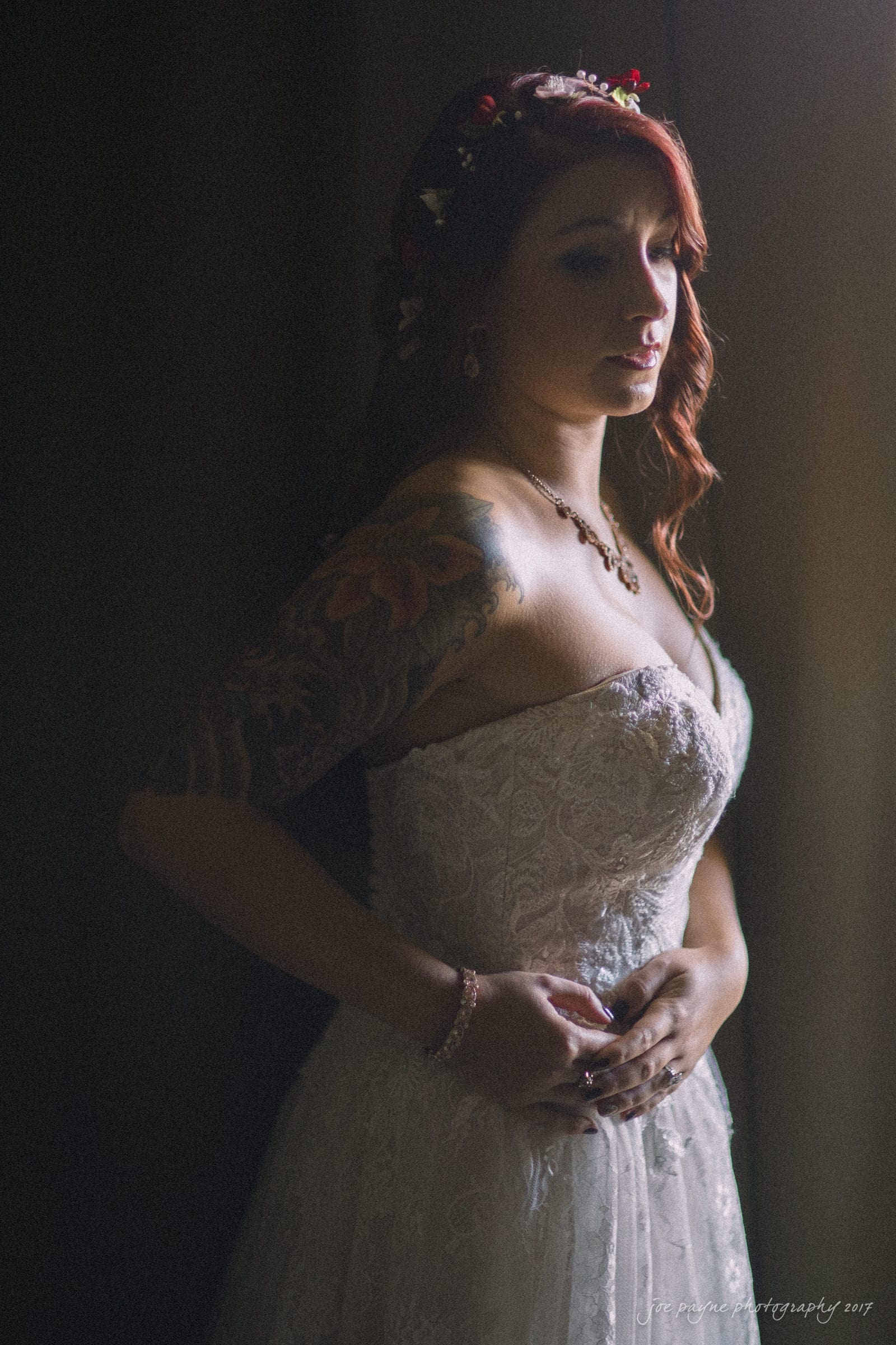 river landing country club wedding - megan & chad Bride in a white lace dress stands in soft lighting, holding her hands at her waist. Her red hair is styled with a floral headpiece, and she has a colorful sleeve tattoo on her arm.