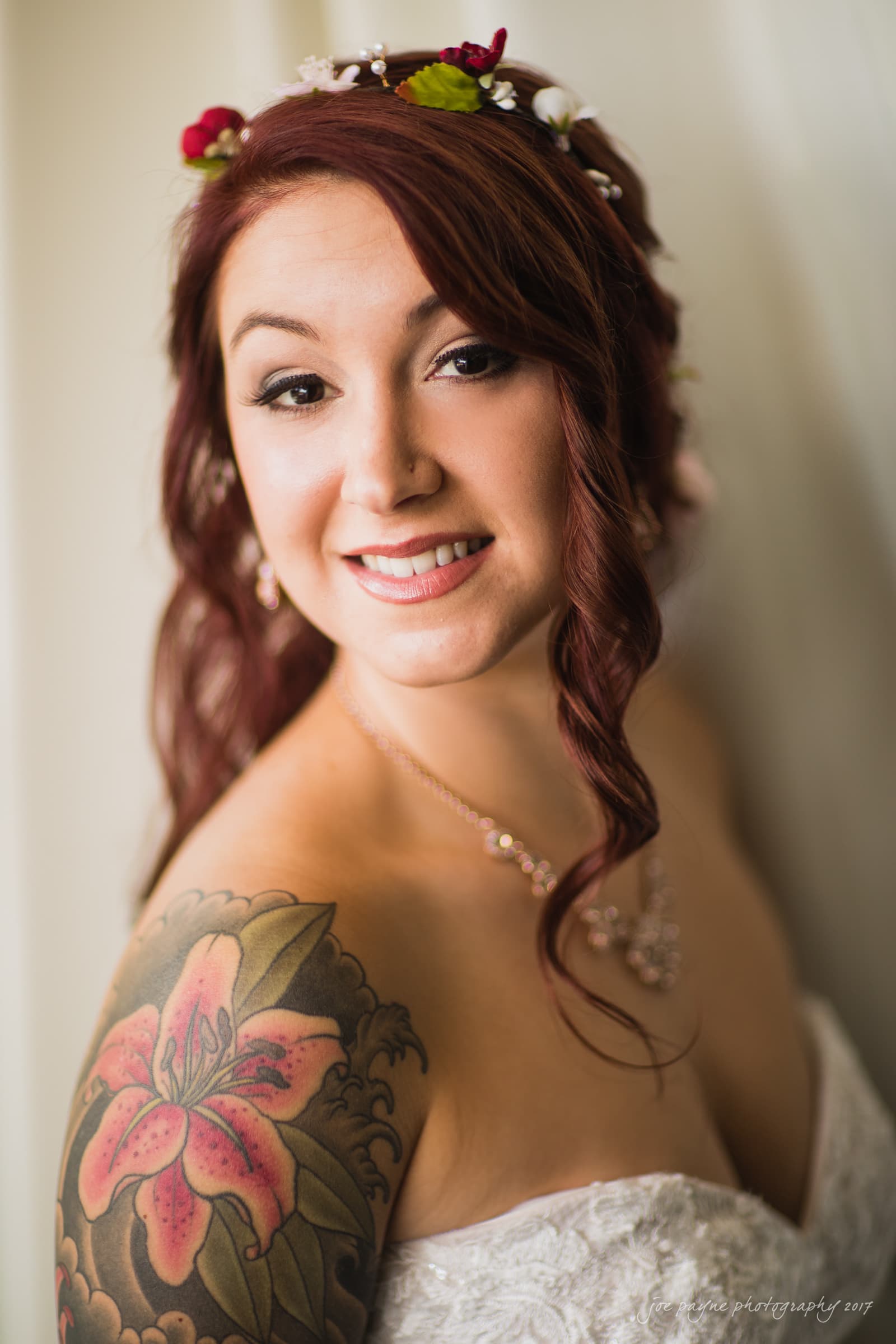 river landing country club wedding - megan & chad A smiling bride with wavy red hair adorned with small flowers poses in a strapless white wedding dress. She has a floral tattoo on her shoulder and is wearing a delicate necklace and earrings.