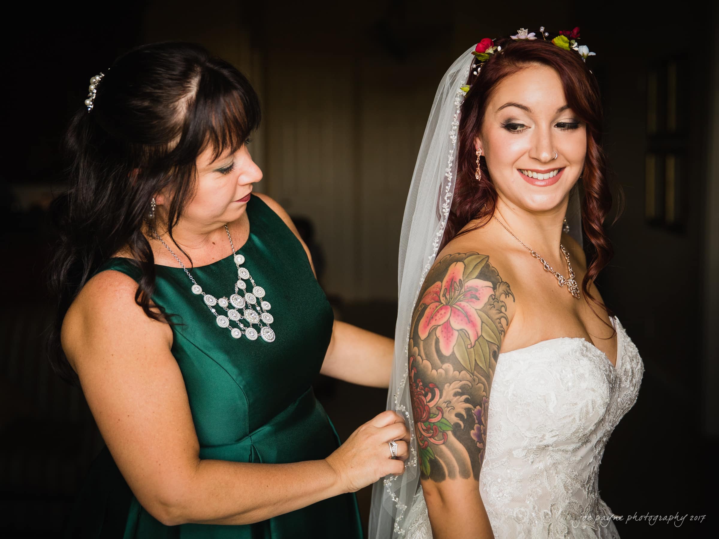 river landing country club wedding - megan & chad A woman wearing a green dress helps a smiling bride with a flower tattoo on her arm adjust her white wedding gown. The bride has a floral crown in her hair, and they are inside a dimly lit room.