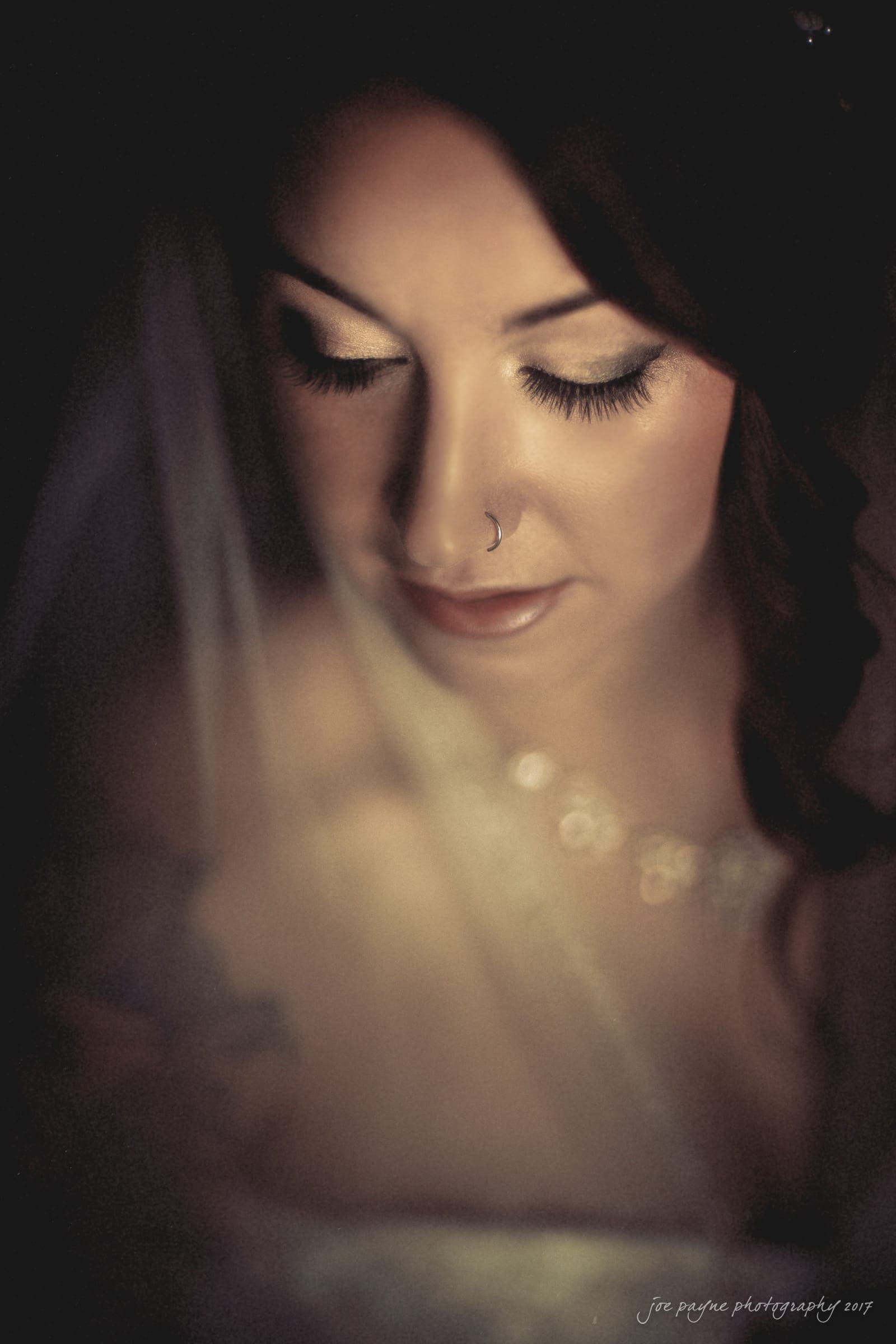 river landing country club wedding - megan & chad A close-up, moody photograph of a bride with soft-focus effects. She is softly lit with eyes closed, wearing a nose ring and a delicate necklace, and draped in a veil. The image has a warm, intimate atmosphere.
