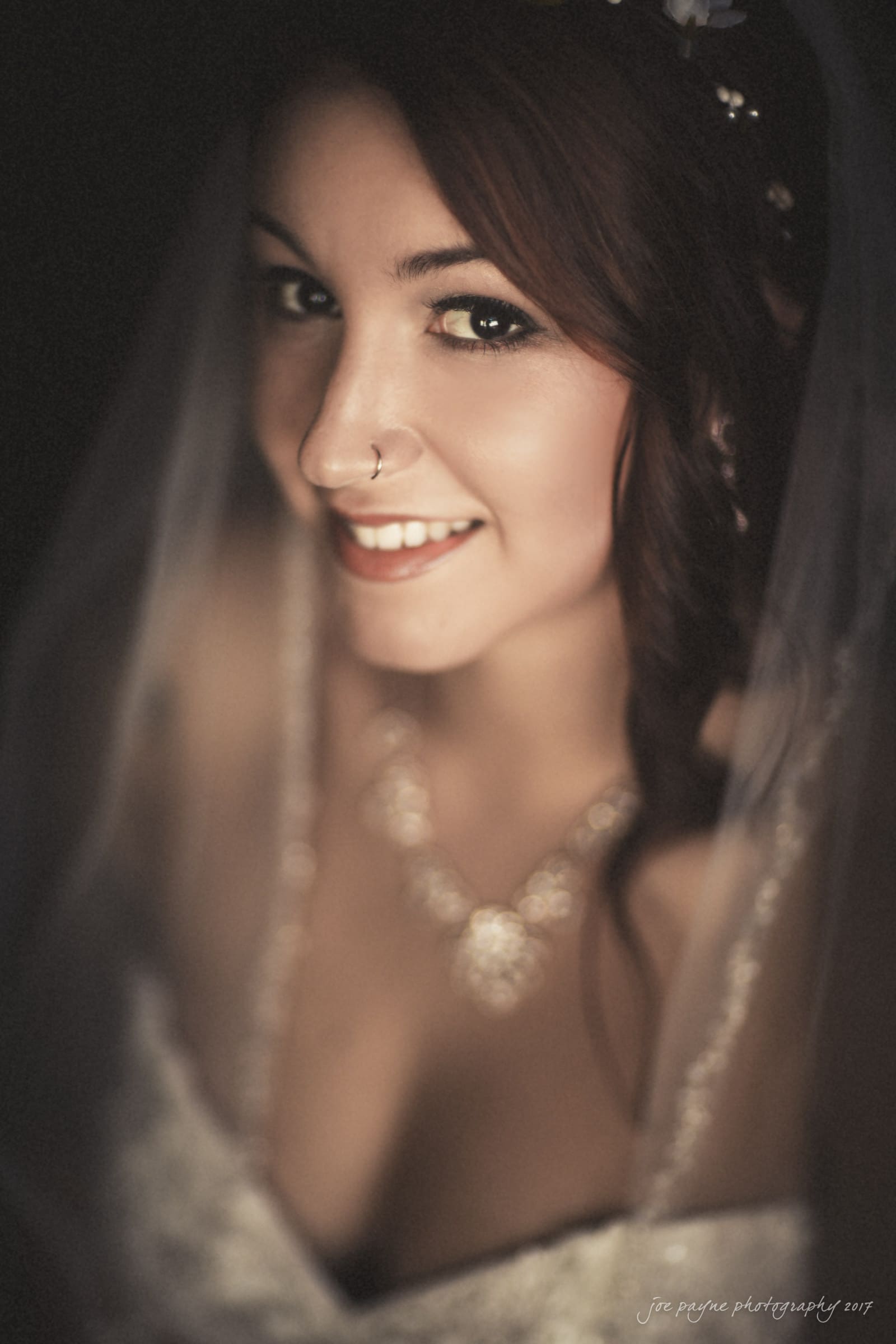 river landing country club wedding - megan & chad A bride with red hair smiles softly. She is wearing a lace wedding dress, a necklace, and a nose ring. The background is dark, emphasizing her features and wedding attire.