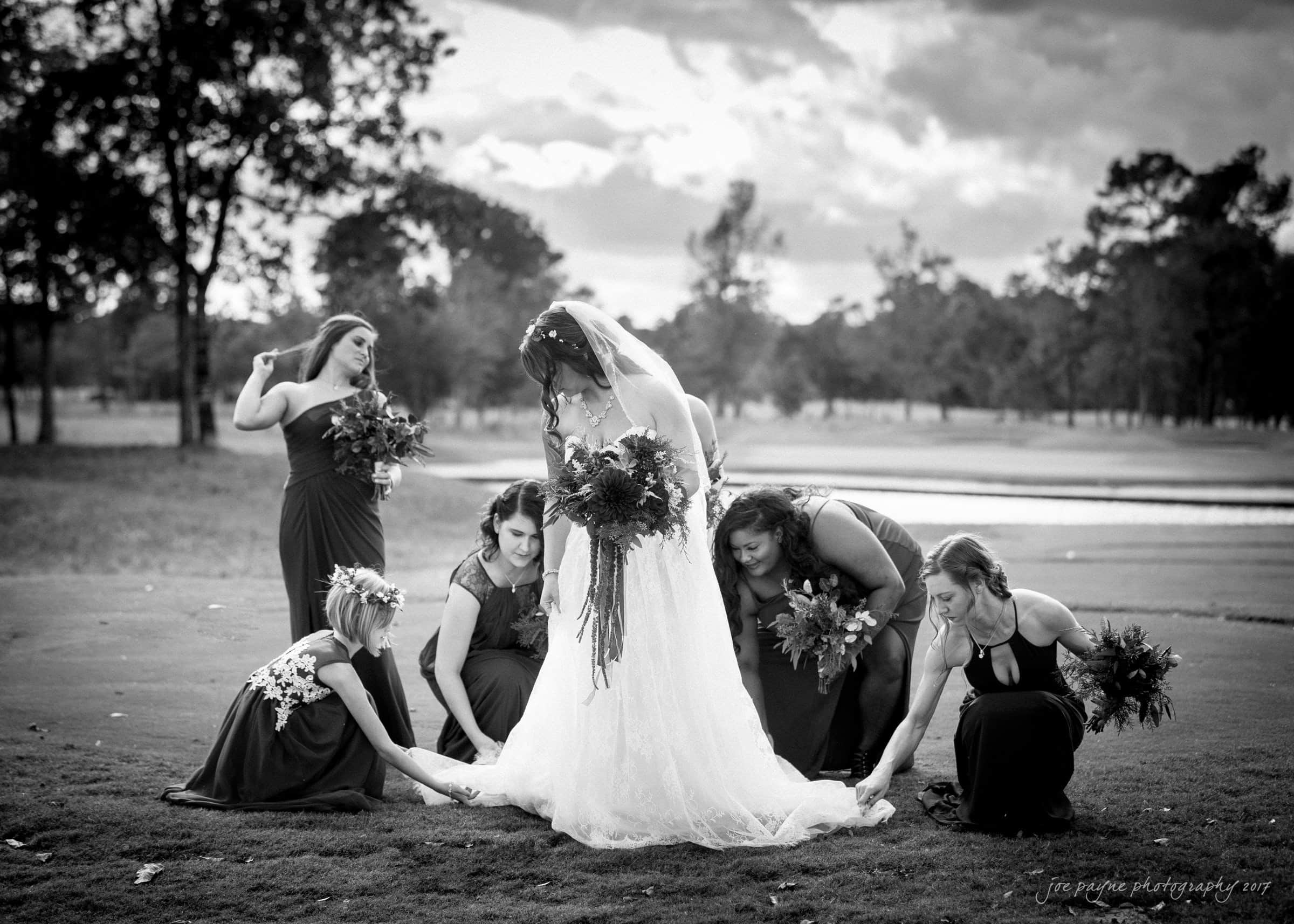 river landing country club wedding - megan & chad A bride in a long dress stands on a lawn, surrounded by bridesmaids adjusting her gown. They hold bouquets, and trees are visible in the background. The scene is in black and white.