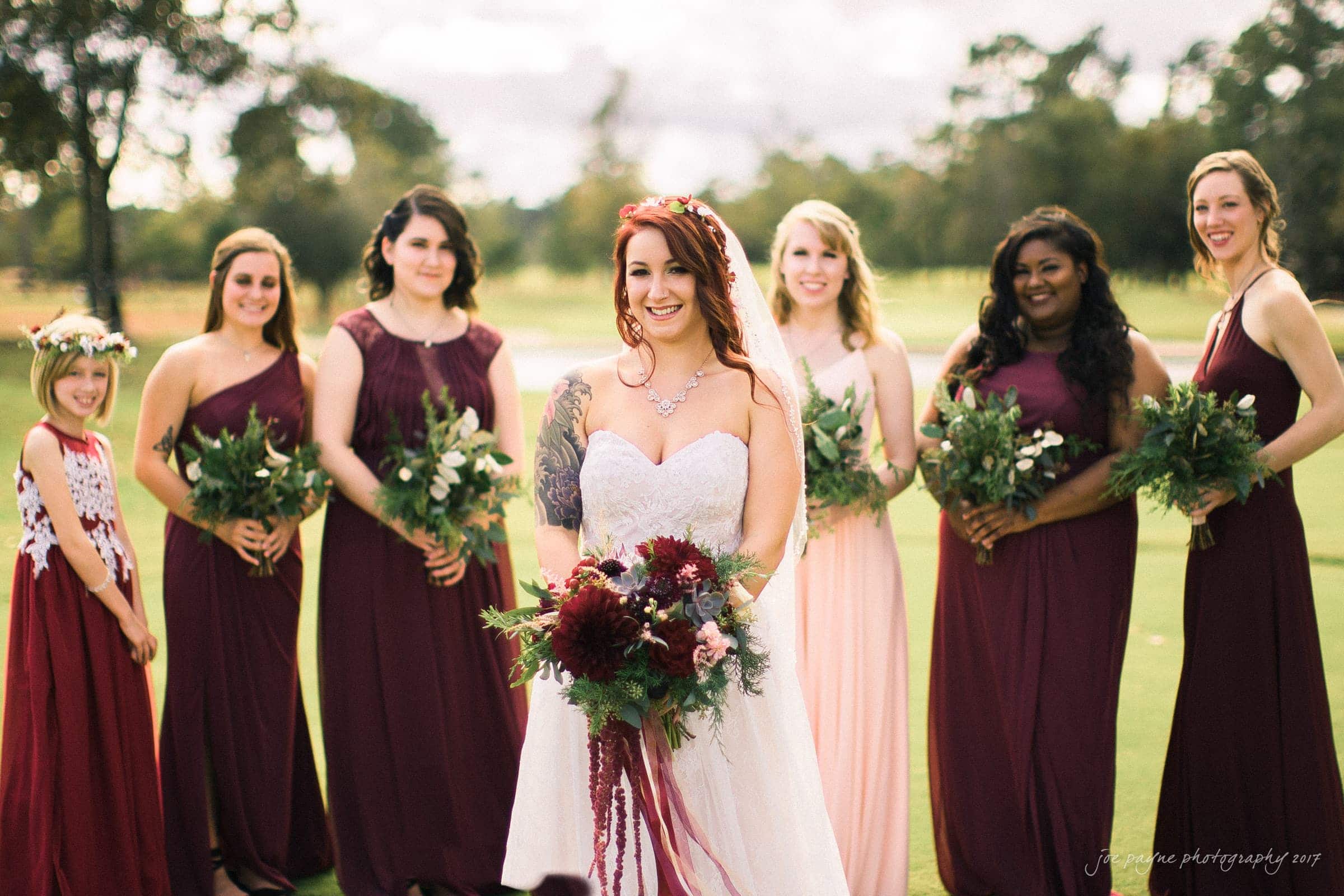 river landing country club wedding - megan & chad A bride in a white gown with a bouquet stands with six bridesmaids wearing maroon and pink dresses. They are smiling outdoors on a grassy area with trees in the background.