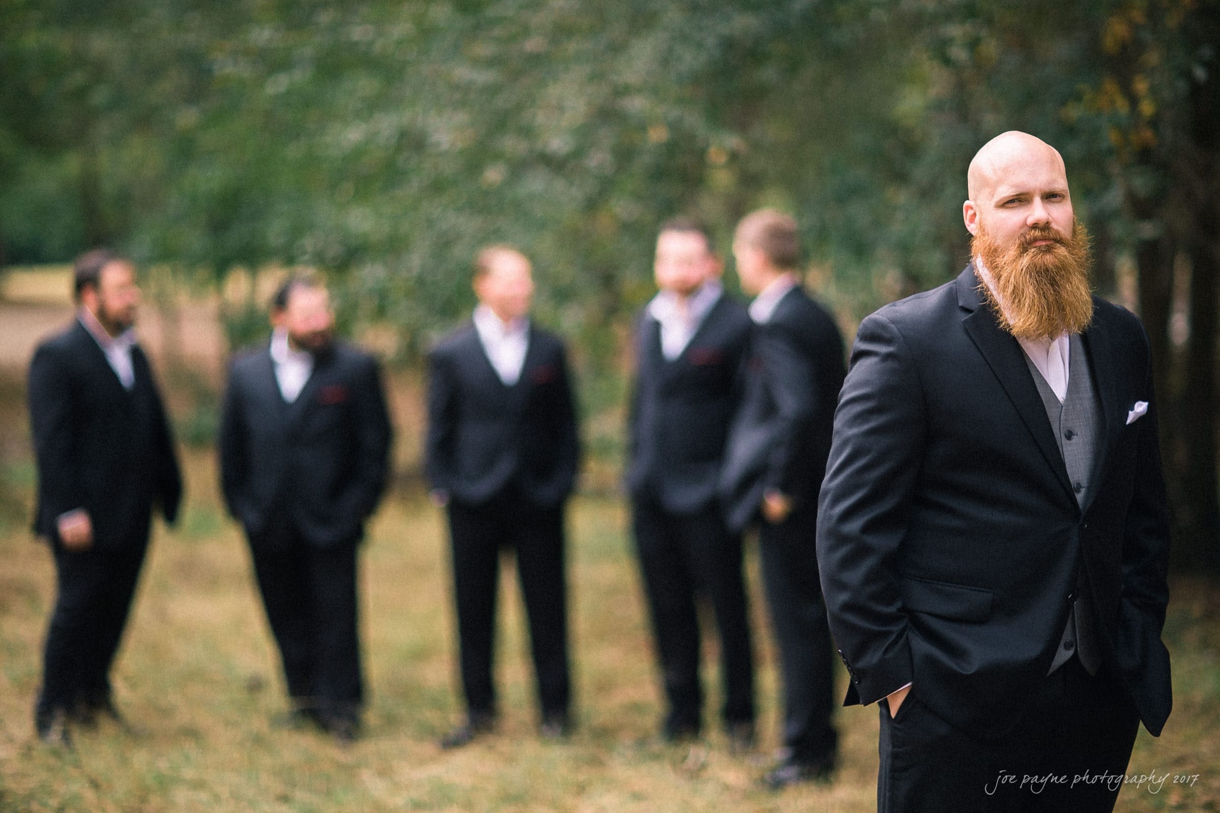 river landing country club wedding - megan & chad A bearded man in a suit stands in focus in the foreground, while four other men in suits are blurred in the background, standing outdoors among trees.