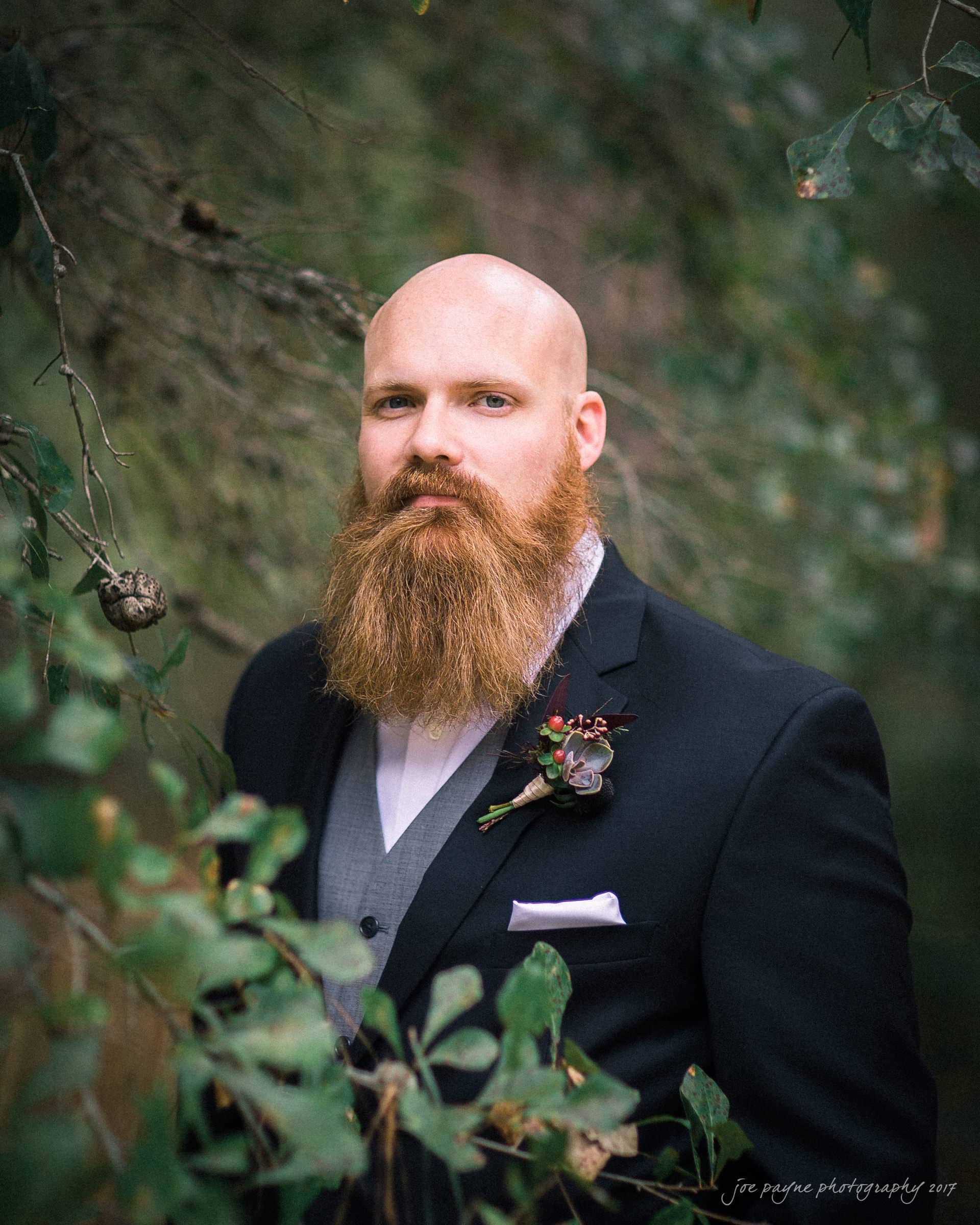 river landing country club wedding - megan & chad A bald man with a full, long beard wearing a black suit and gray vest stands among lush greenery. He has a boutonniere on his lapel and looks directly at the camera, set against a blurred leafy background.
