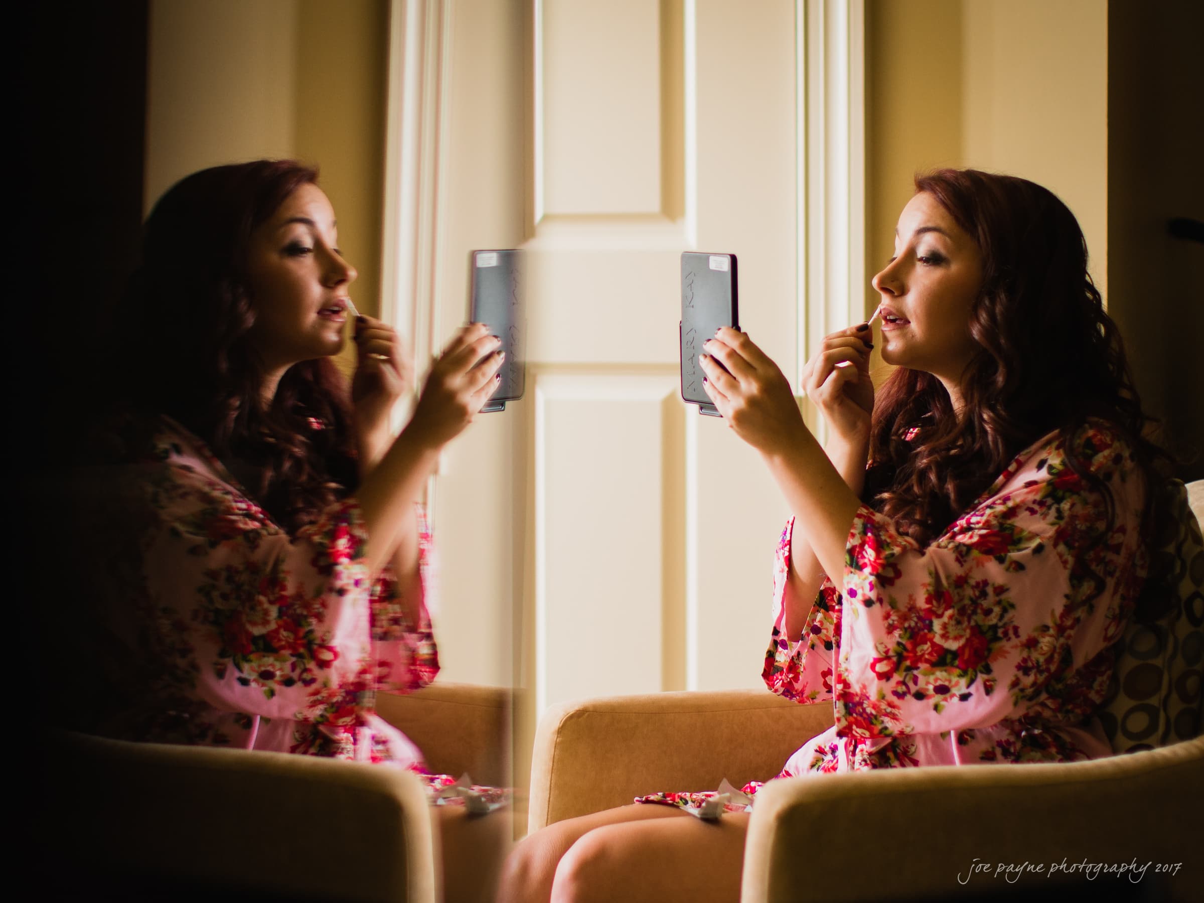 river landing country club wedding - megan & chad A woman with long hair applies lipstick while looking into a handheld mirror. She is seated on a chair and wears a floral robe. Her reflection is visible on a glass surface beside her. The setting appears warm and well-lit.