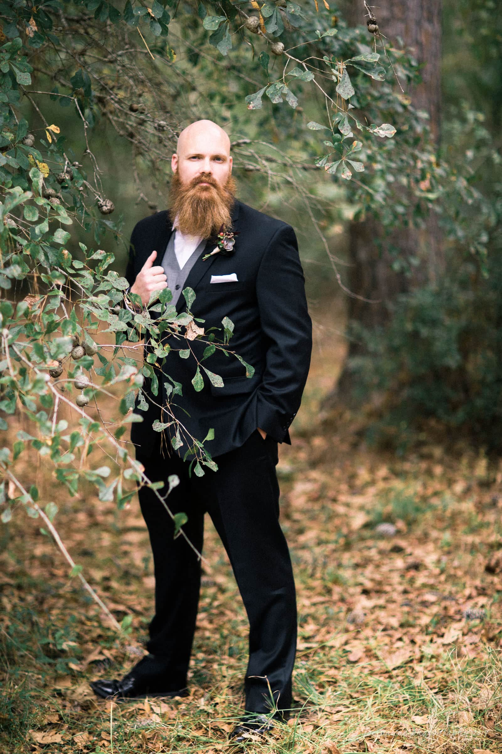 river landing country club wedding - megan & chad A bearded man in a black suit stands confidently outdoors among trees and foliage. He poses with one hand in his pocket and the other adjusting his lapel, surrounded by fallen leaves and green branches.