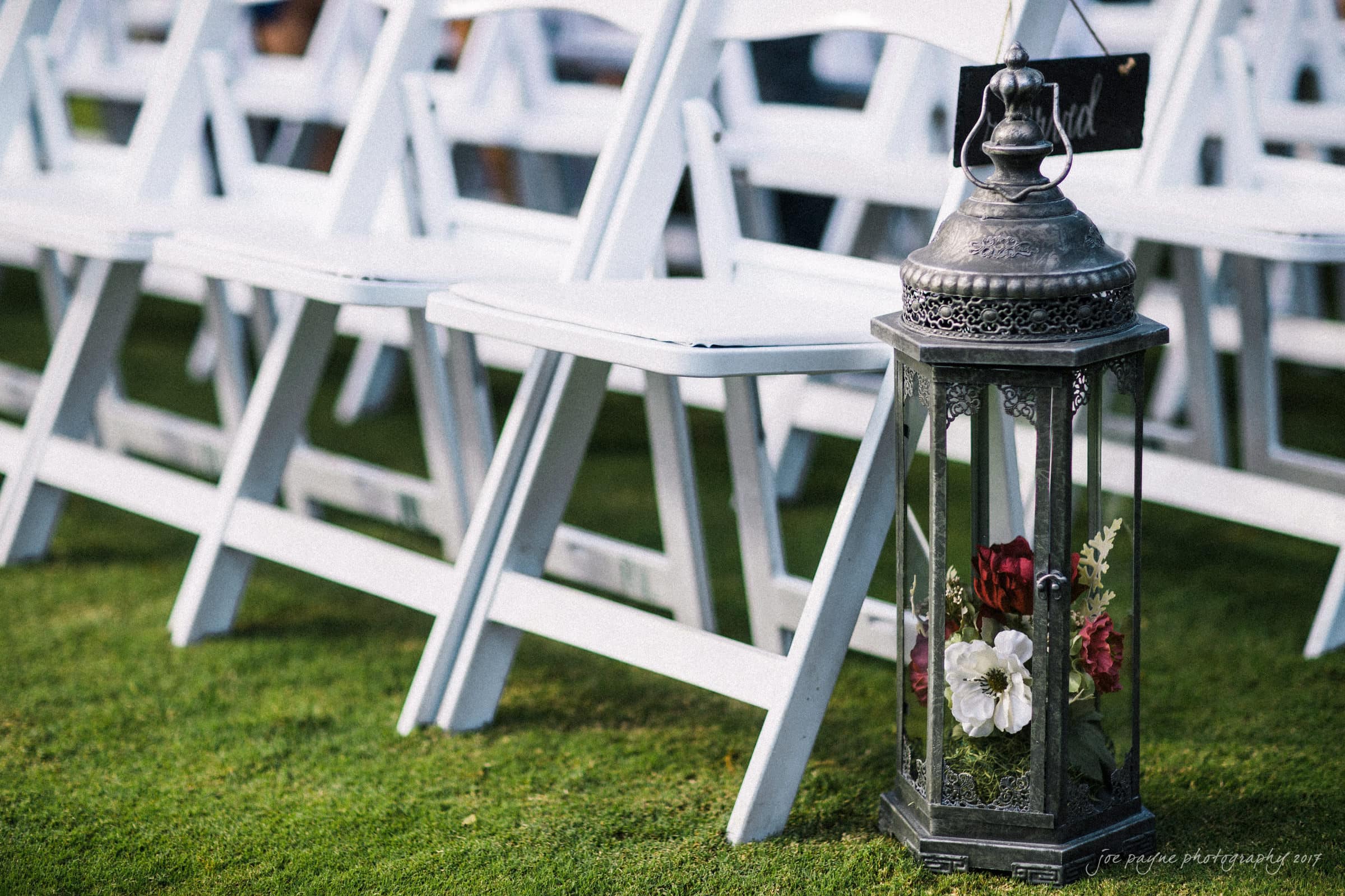 river landing country club wedding - megan & chad A decorative lantern with red and white flowers inside rests on grass beside white folding chairs arranged for an outdoor event.