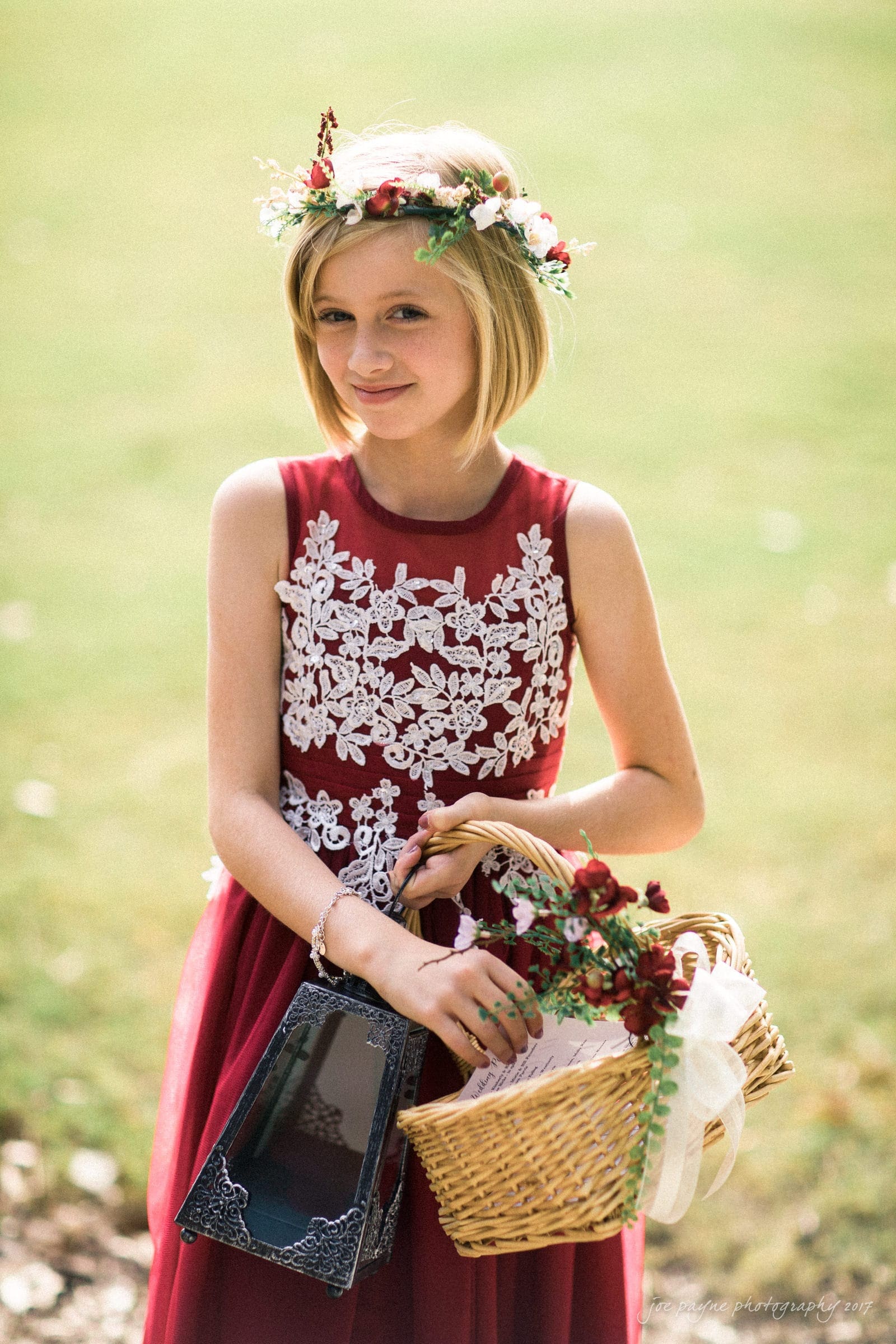 river landing country club wedding - megan & chad A young girl in a red, floral-lace dress and flower crown stands outdoors, holding a basket filled with flowers and a black bag. She is smiling softly in a natural setting with green grass in the background.