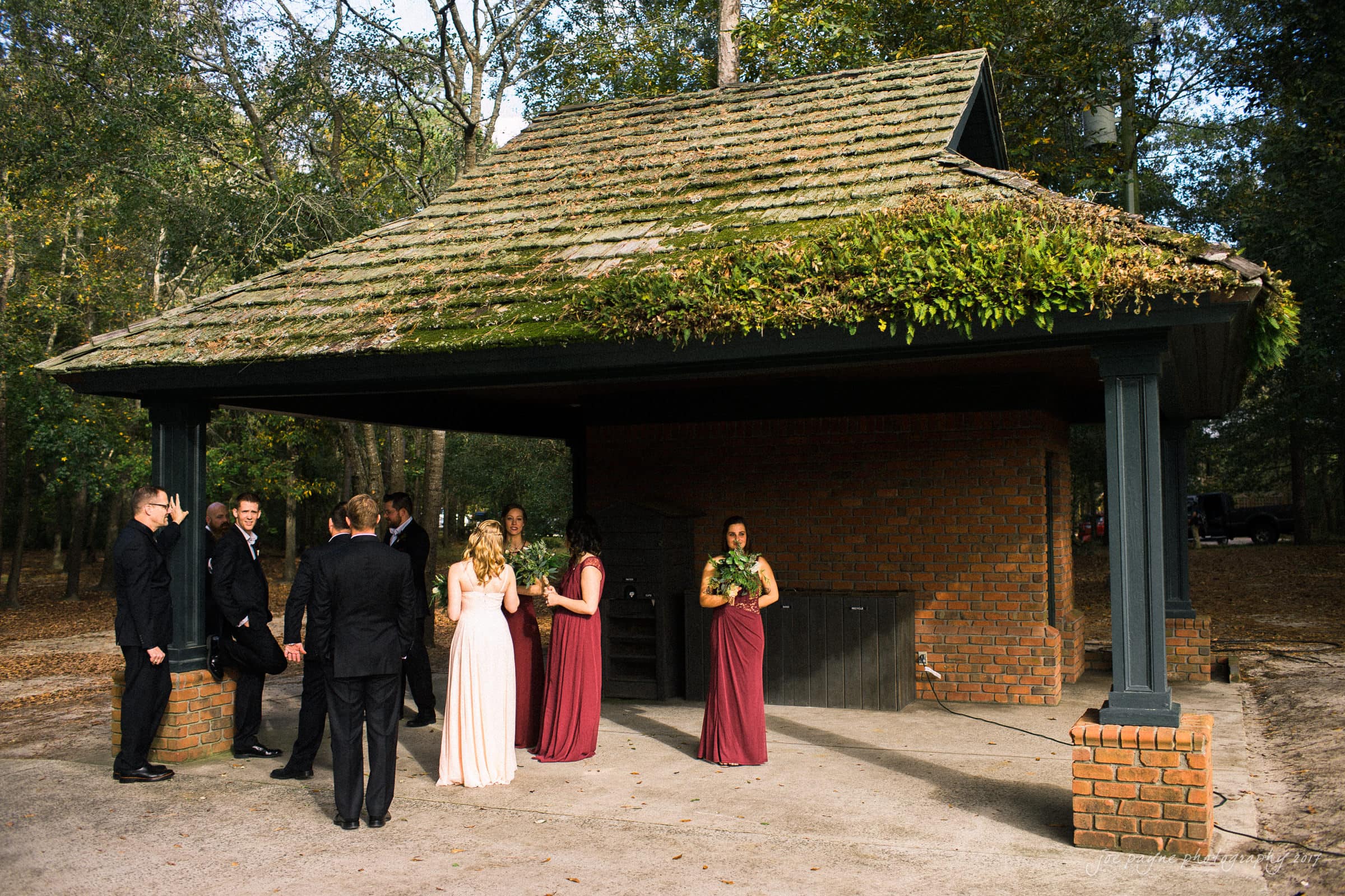 river landing country club wedding - megan & chad A group of people in formal attire stand outside, under a small open shelter with a shingled roof, surrounded by trees. Some women hold bouquets, suggesting a bridal party gathering in a park or outdoor setting.