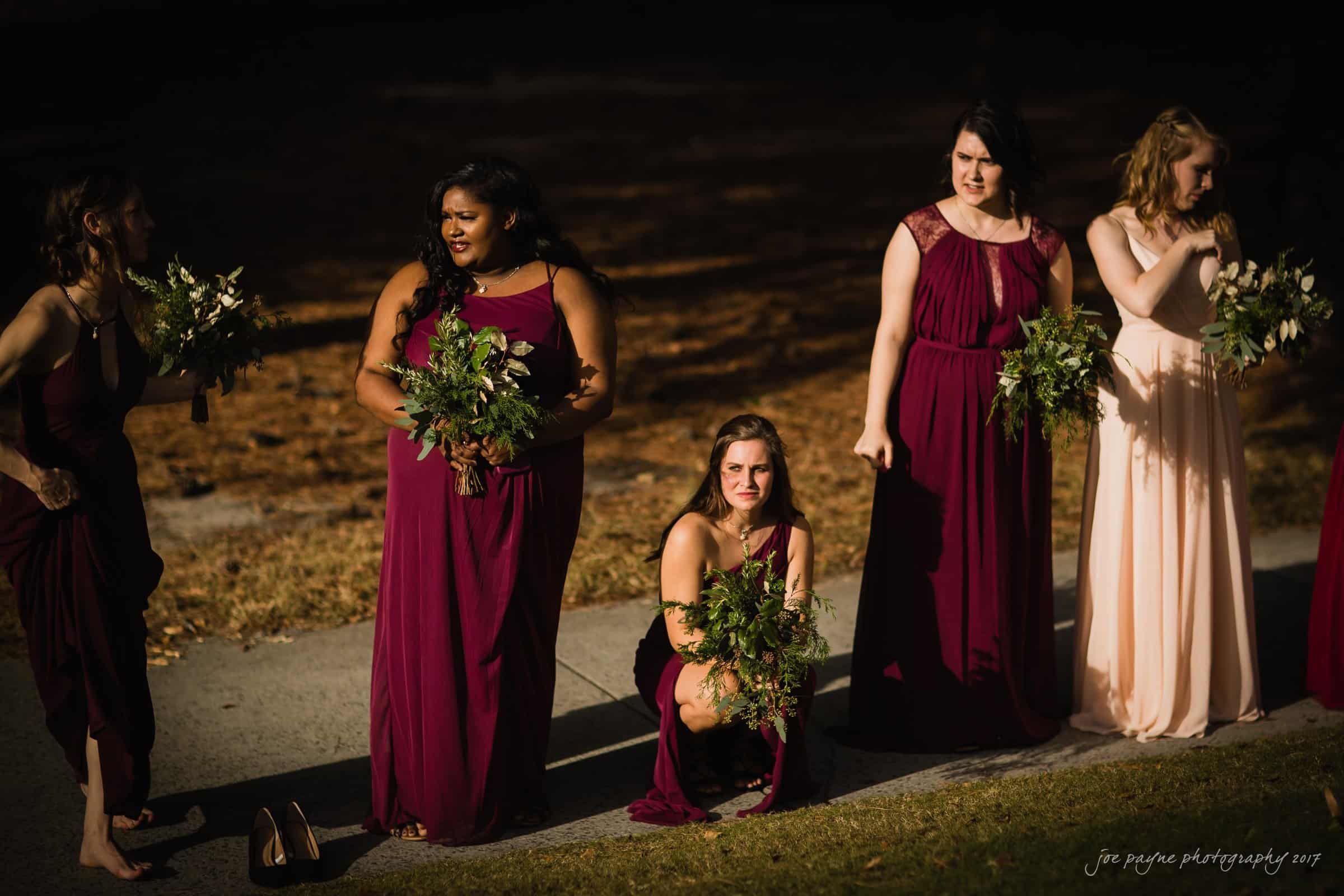 river landing country club wedding - megan & chad A group of five bridesmaids in maroon and blush dresses, holding bouquets, stand on a sunlit path with trees in the background. One bridesmaid crouches down, smiling. The scene is lively and cheerful.