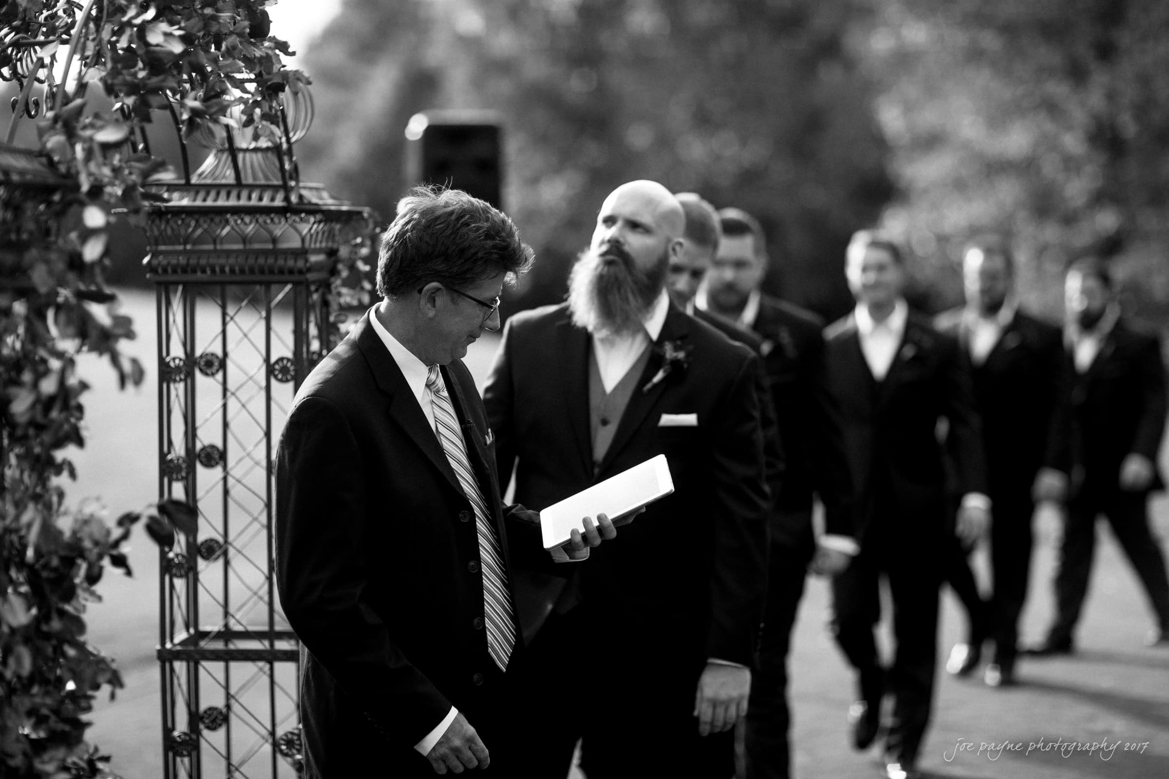 river landing country club wedding - megan & chad A group of men in suits walking outdoors. One with a beard stands prominently in the foreground, while another man holds a book and looks down. The scene is black and white, with trees and an outdoor structure in the background.