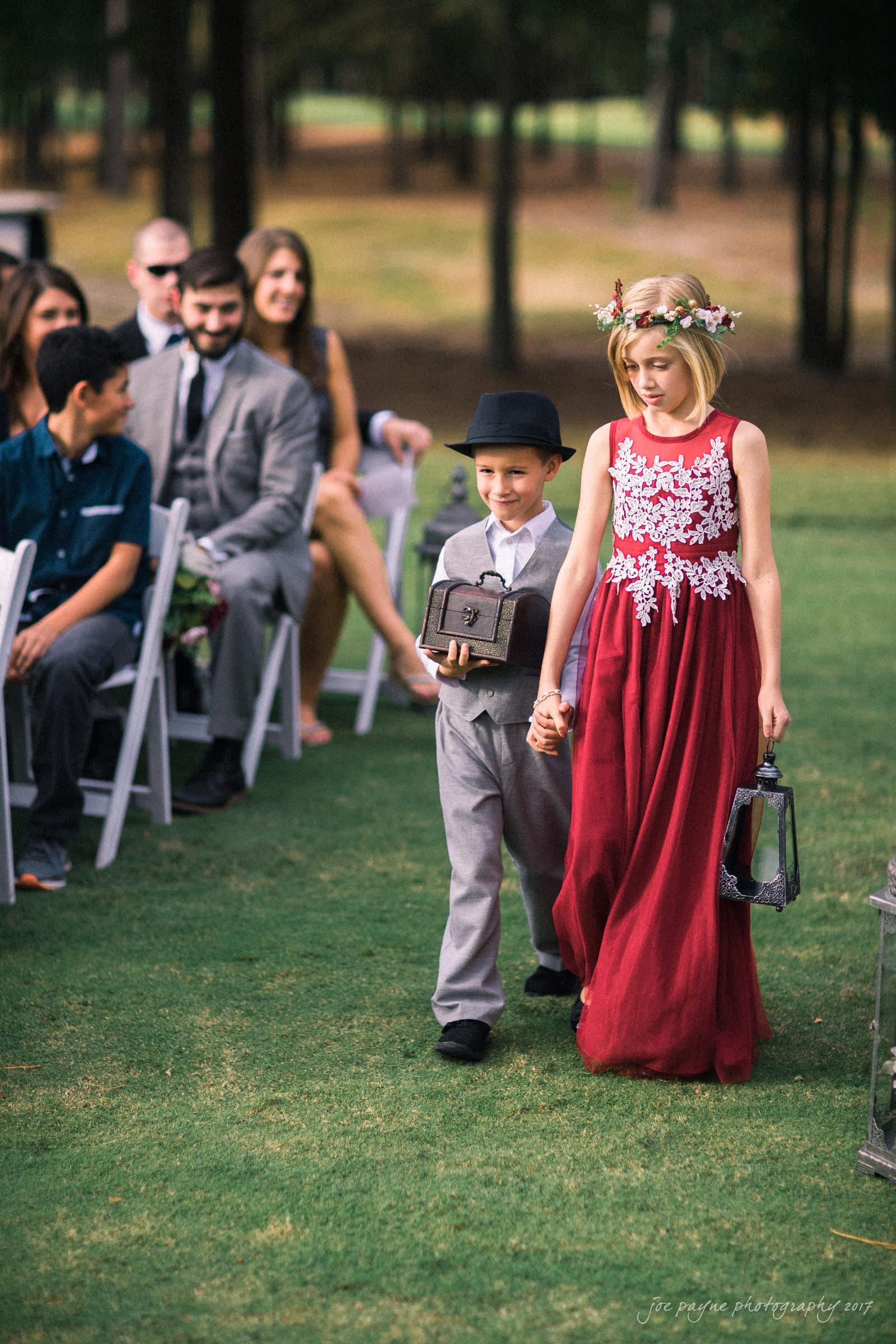 river landing country club wedding - megan & chad A young girl in a red dress and flower crown walks hand in hand with a boy in a gray suit and hat. They are on a grassy aisle, surrounded by seated people at an outdoor event. The boy carries a small wooden box.