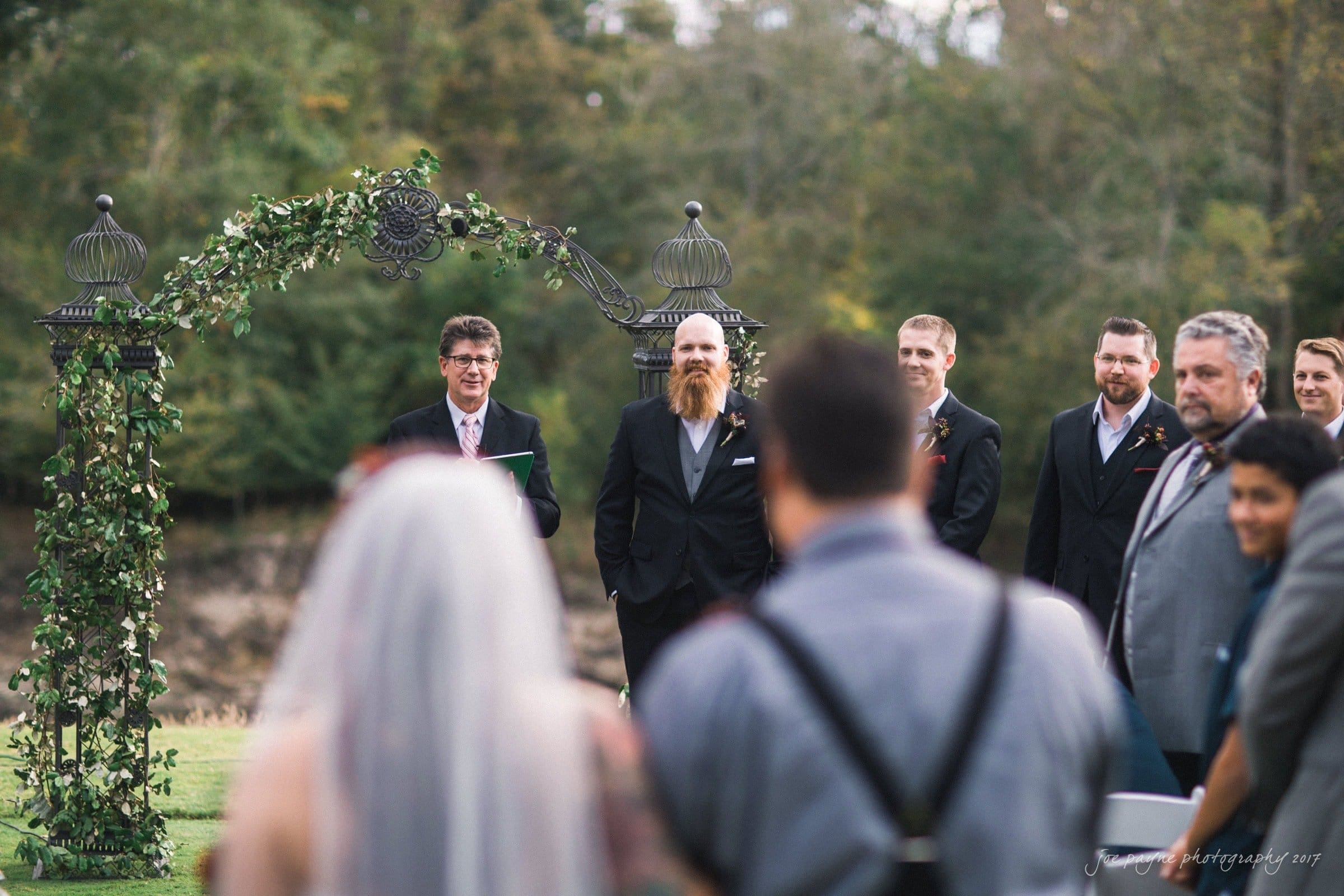 river landing country club wedding - megan & chad A wedding ceremony outdoors with a leafy arch. A bride and groom stand facing an officiant. Several guests, including men in suits, are visible. The background features trees and grass, creating a serene and natural setting.