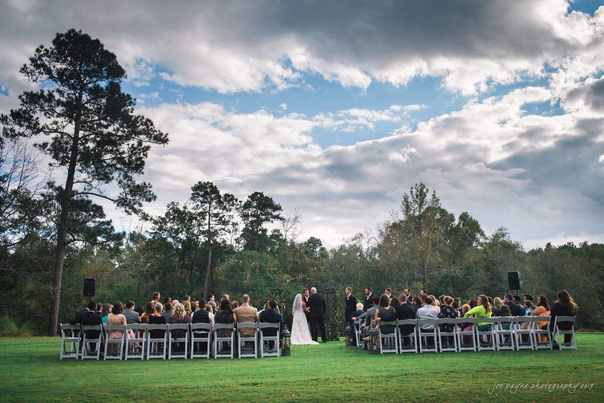 river landing country club wedding - megan & chad A couple is getting married outdoors under a partly cloudy sky. Guests are seated on white chairs, and a forested area surrounds the scene. The setting is serene, with a mix of greenery and open sky.