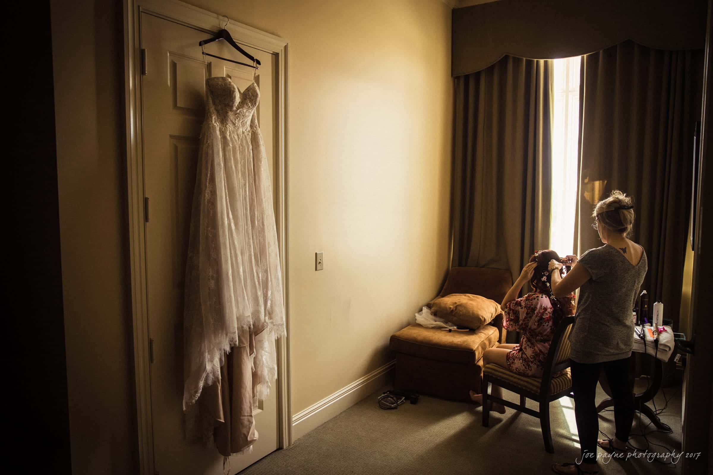 river landing country club wedding - megan & chad A wedding dress hangs on a door in a dimly lit room. A woman sits in a chair getting her hair styled by a hairstylist. A small dog naps on a cushion beside them. Curtains partially cover a window, casting soft light into the space.