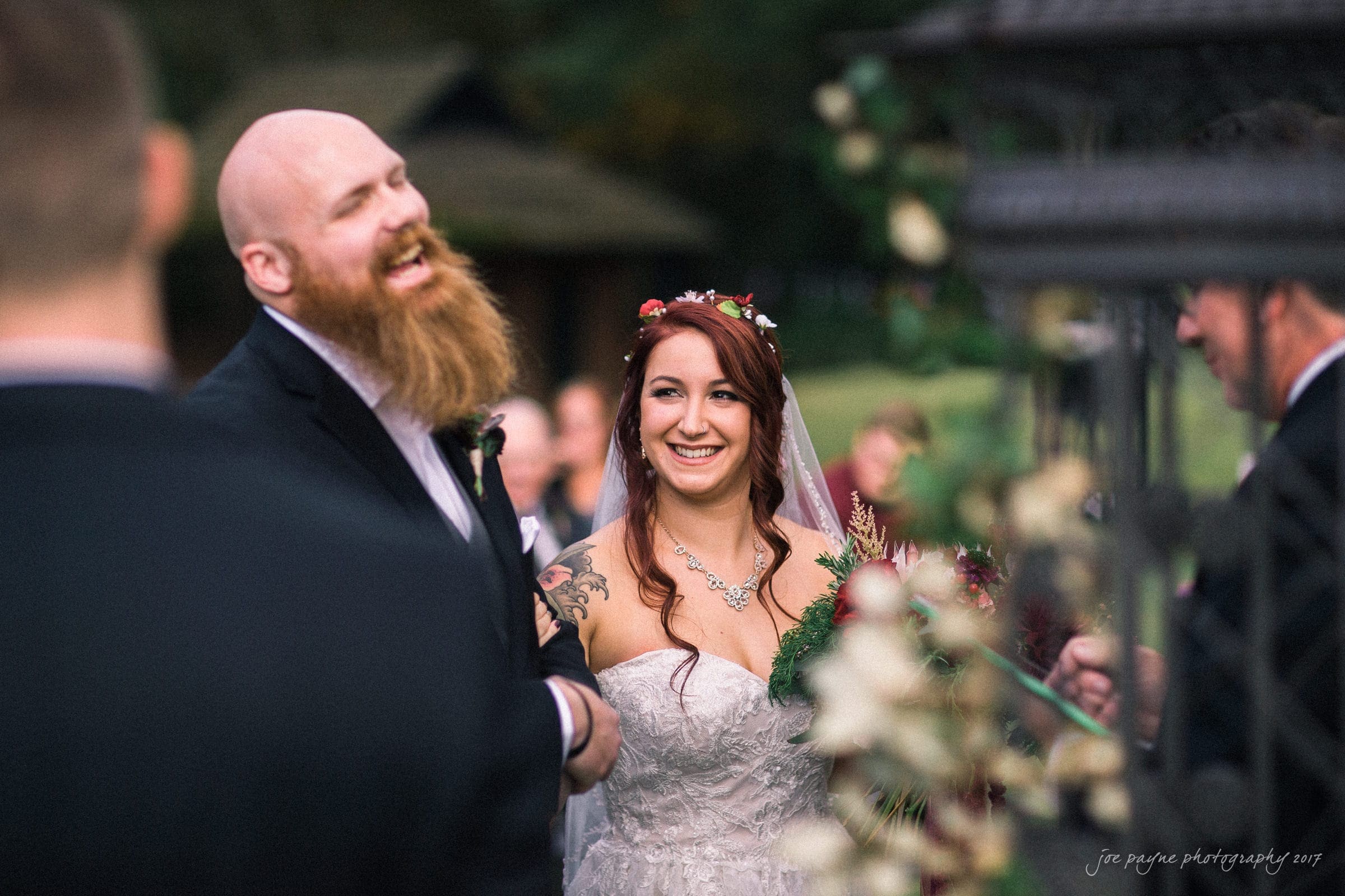 river landing country club wedding - megan & chad A bride and groom laugh joyfully during their outdoor wedding ceremony. The bride wears a floral headpiece and a lace dress, while the groom has a bald head and a long, reddish beard. They are surrounded by guests and a floral backdrop.