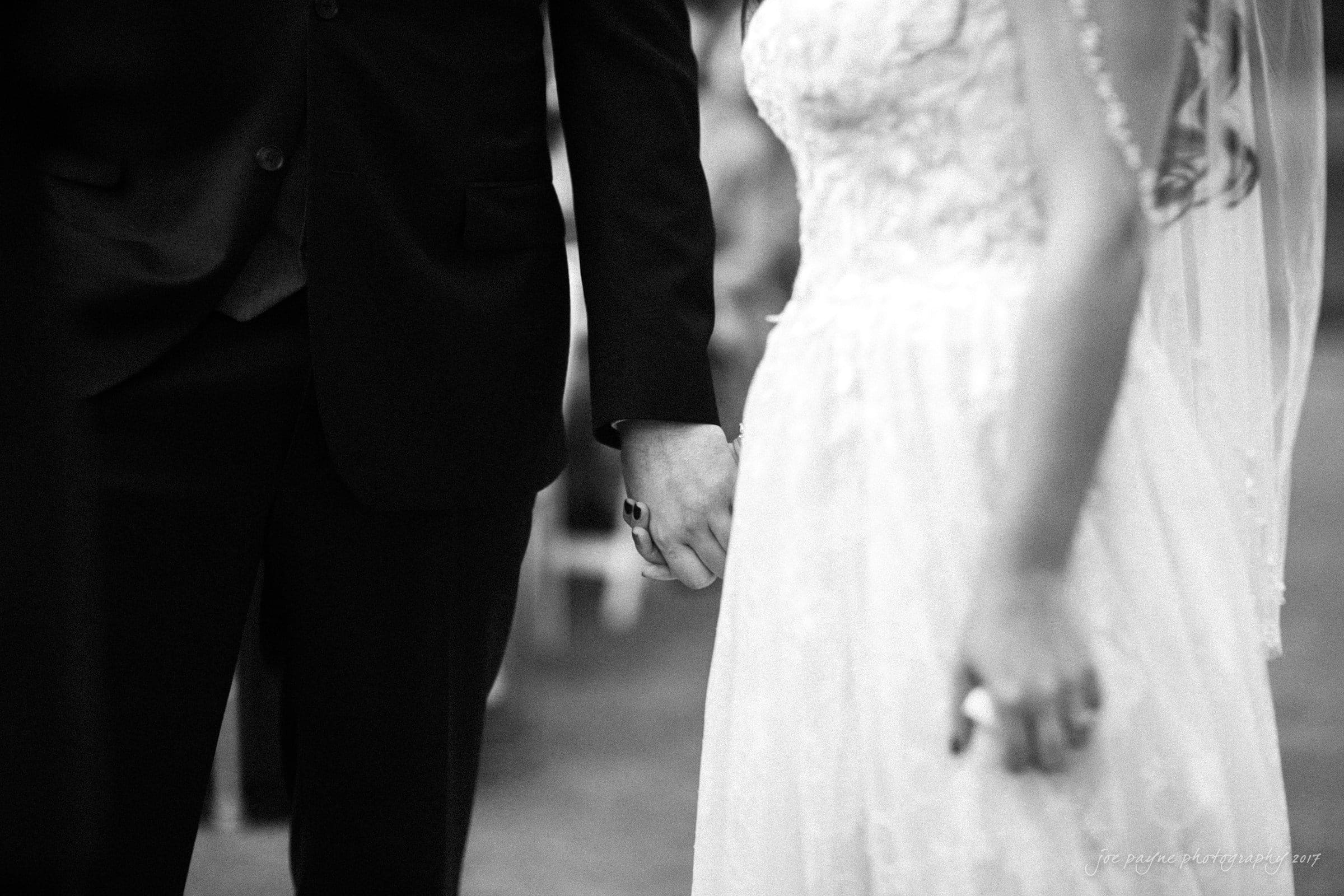 river landing country club wedding - megan & chad A black and white photo capturing a bride and groom holding hands. The bride is wearing a lace wedding gown and veil, while the groom is in a dark suit. They stand closely together, symbolizing unity and love.