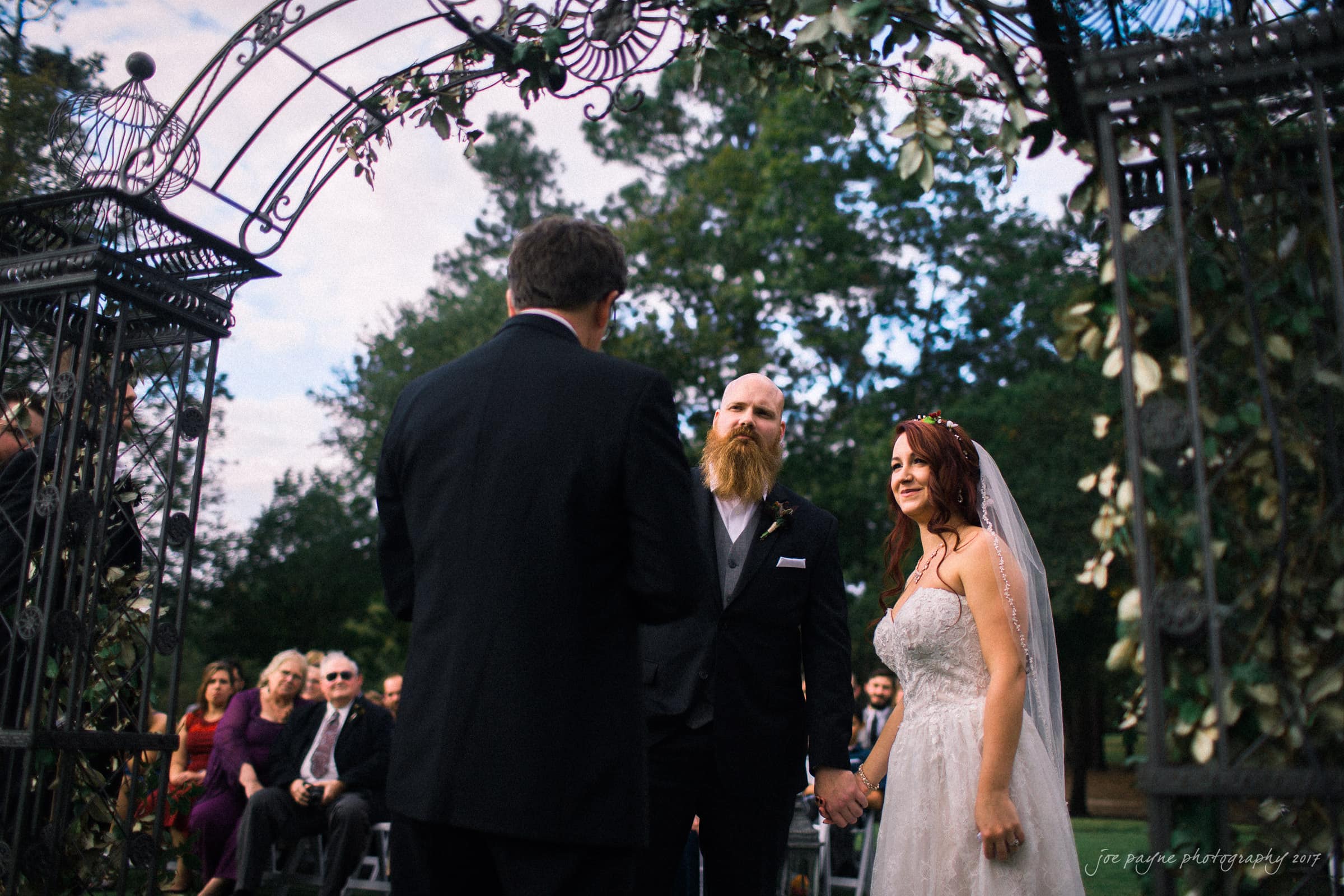 river landing country club wedding - megan & chad A bride and groom stand under a decorative arch during an outdoor wedding ceremony. The groom wears a suit, and the bride is in a white dress with a veil. Guests are seated in the background under a clear sky.