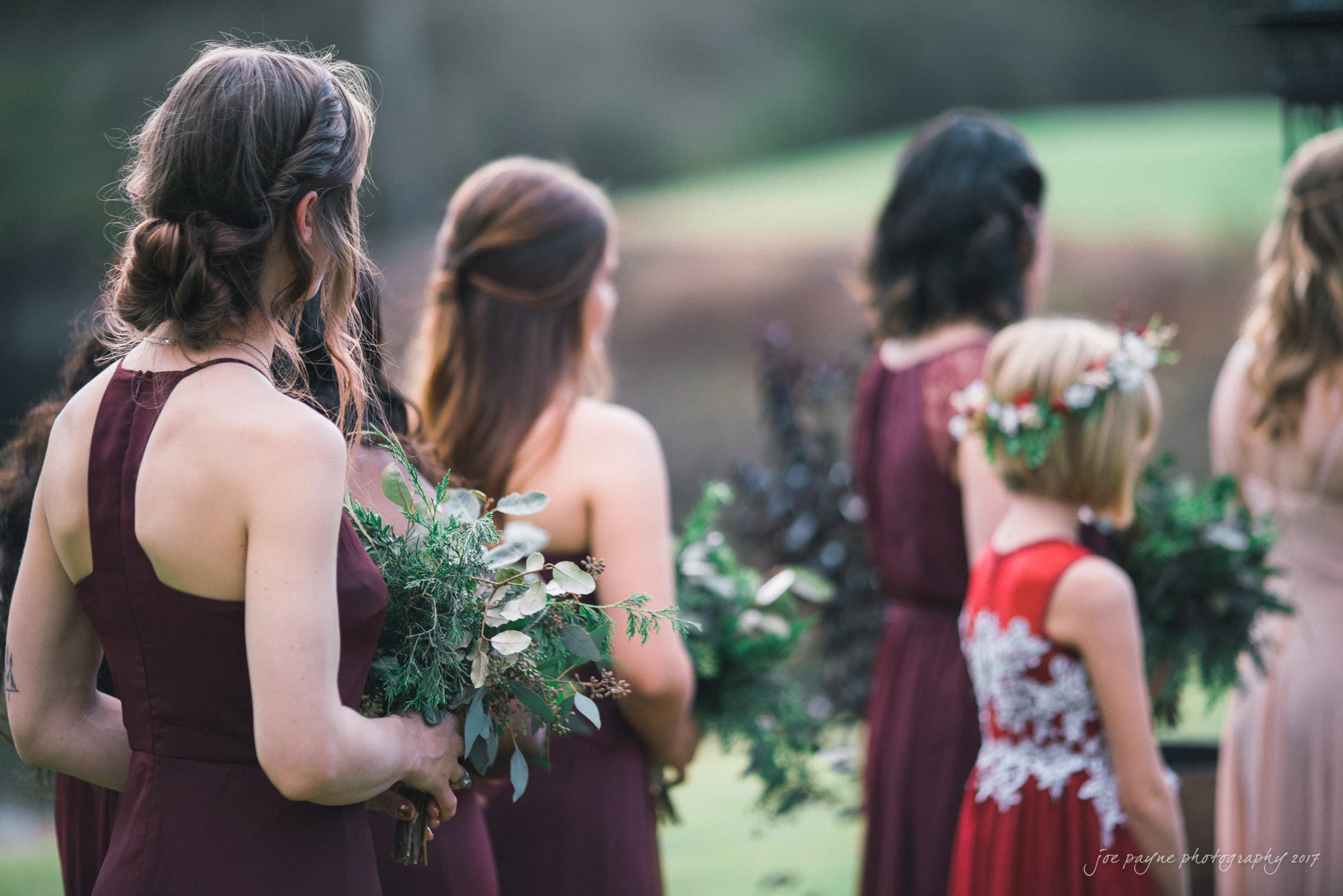 river landing country club wedding - megan & chad A group of bridesmaids in burgundy dresses holding bouquets stand outdoors. A young girl in a lace dress with a flower crown is in the foreground. The background features greenery and blurred landscape.