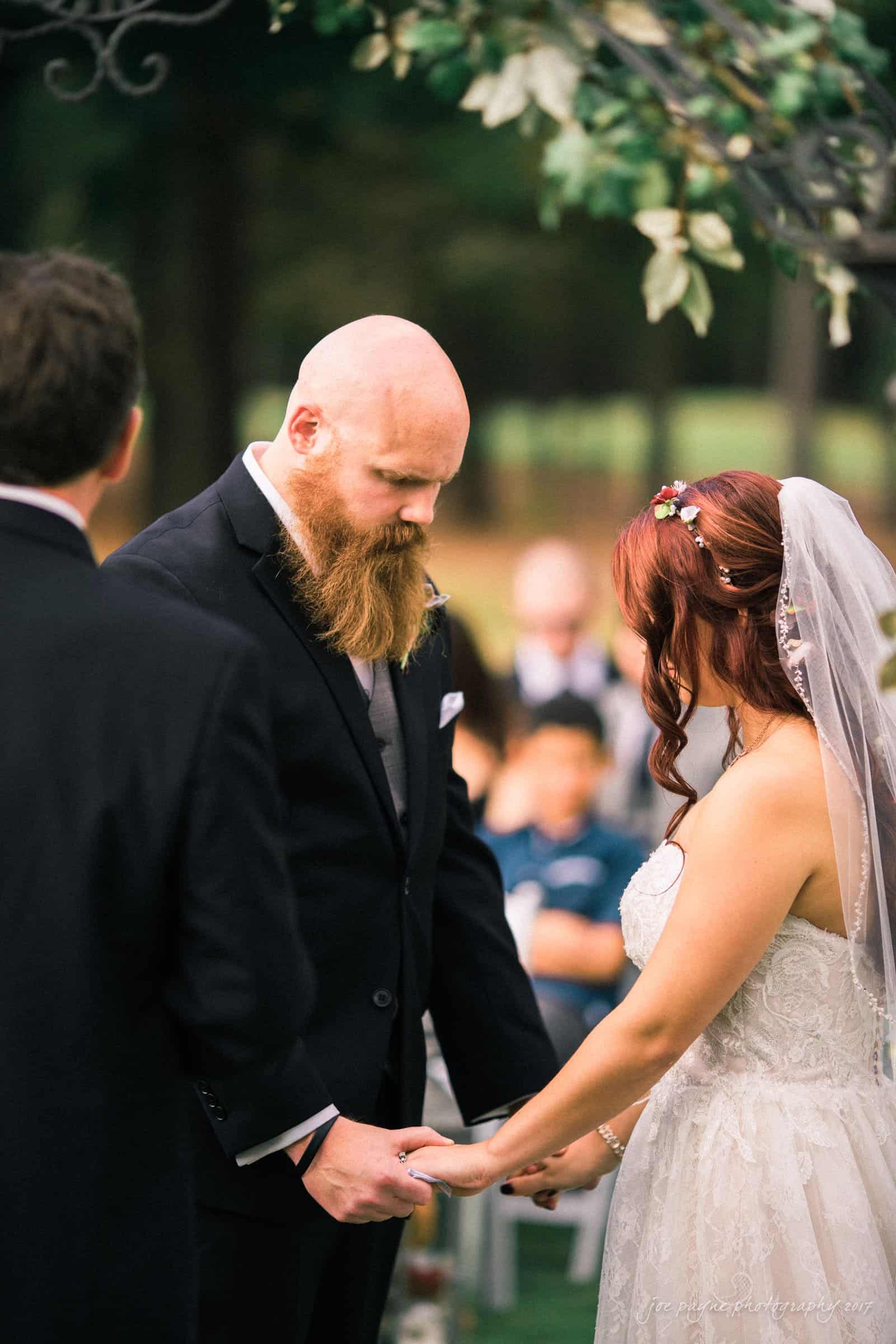 river landing country club wedding - megan & chad A couple stands outdoors, holding hands during a wedding ceremony. The groom has a beard and wears a black suit, while the bride has a veil and wears a white dress. They appear focused and solemn, with greenery visible in the background.