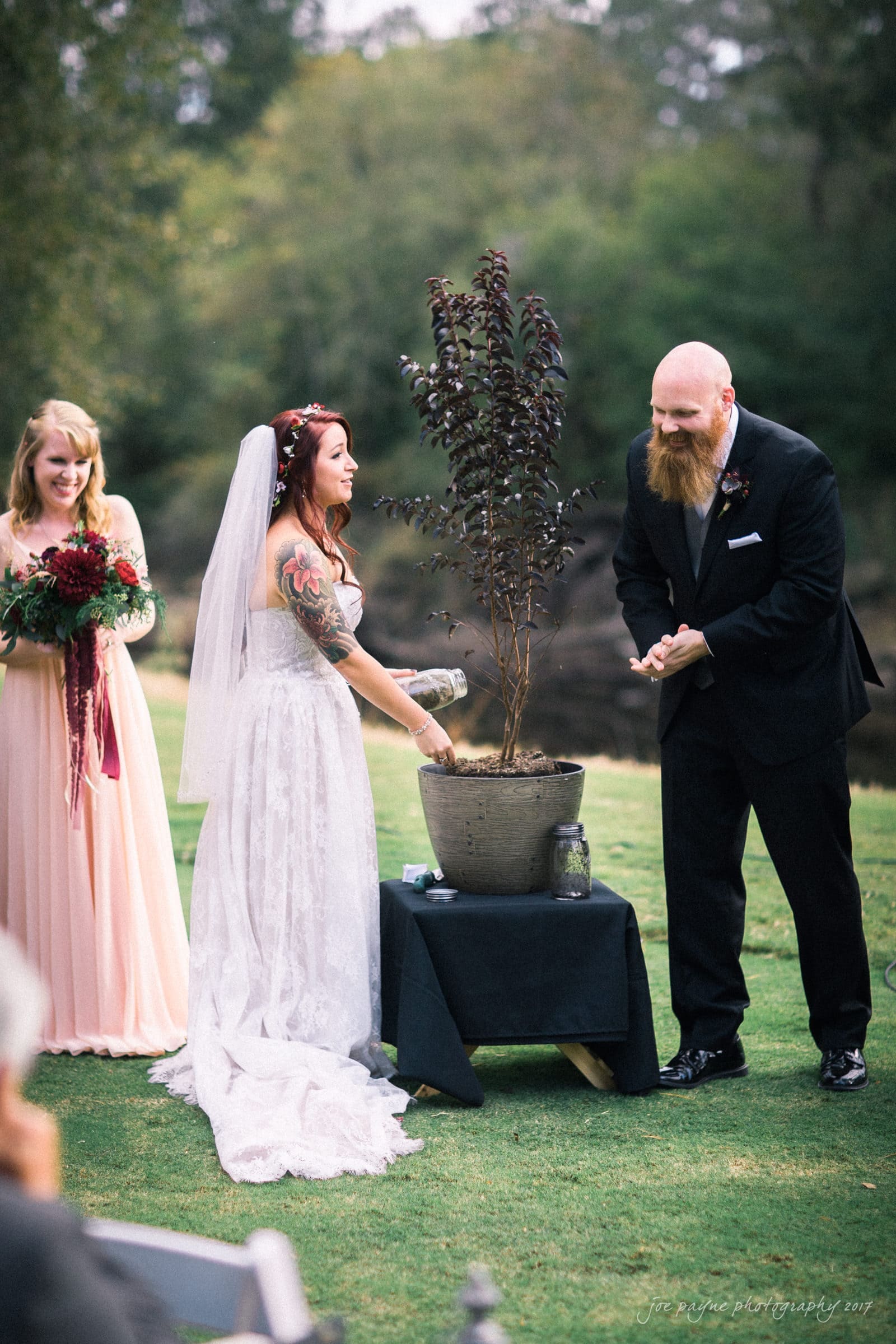 river landing country club wedding - megan & chad A bride and groom participate in a wedding unity ritual with a potted plant outdoors. The bride pours water into the pot while the groom watches. A bridesmaid, holding a bouquet, stands nearby, smiling. Grass and trees are in the background.