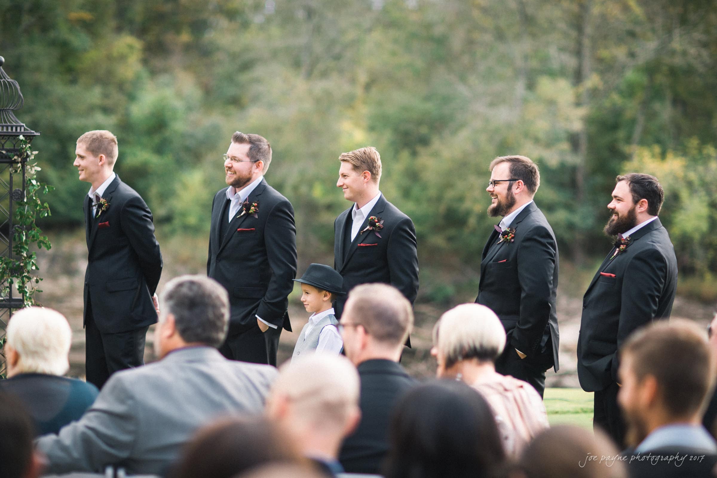 river landing country club wedding - megan & chad A group of five groomsmen in black suits stand in a row outside, with trees in the background. A young boy in a hat stands beside them. The audience is seated in the foreground, partially visible.