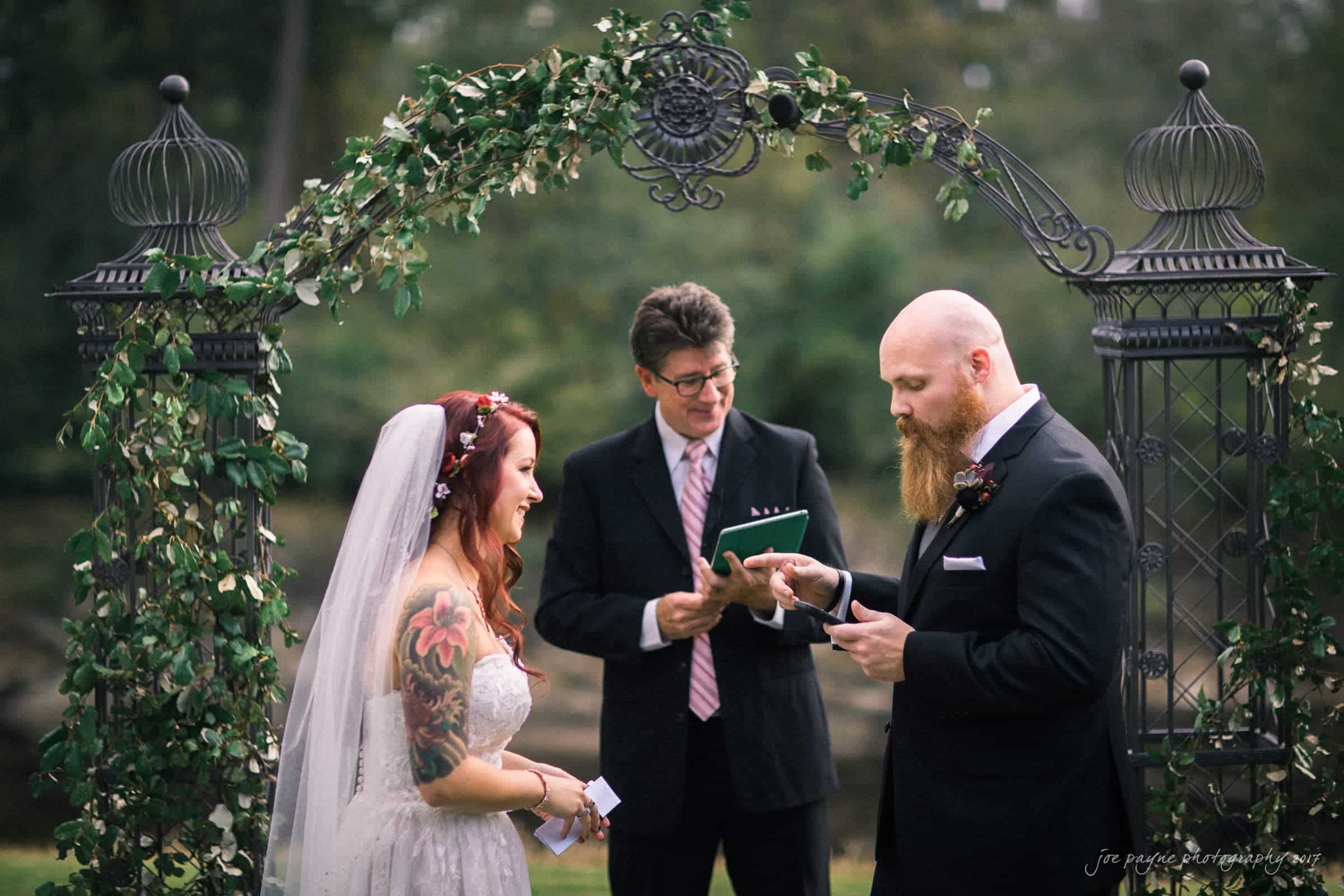 river landing country club wedding - megan & chad A bride and groom stand facing each other under a decorative archway adorned with greenery. The groom, bearded and formally dressed, reads from a card. The bride, tattooed and wearing a veil, smiles. A person officiating stands behind them.