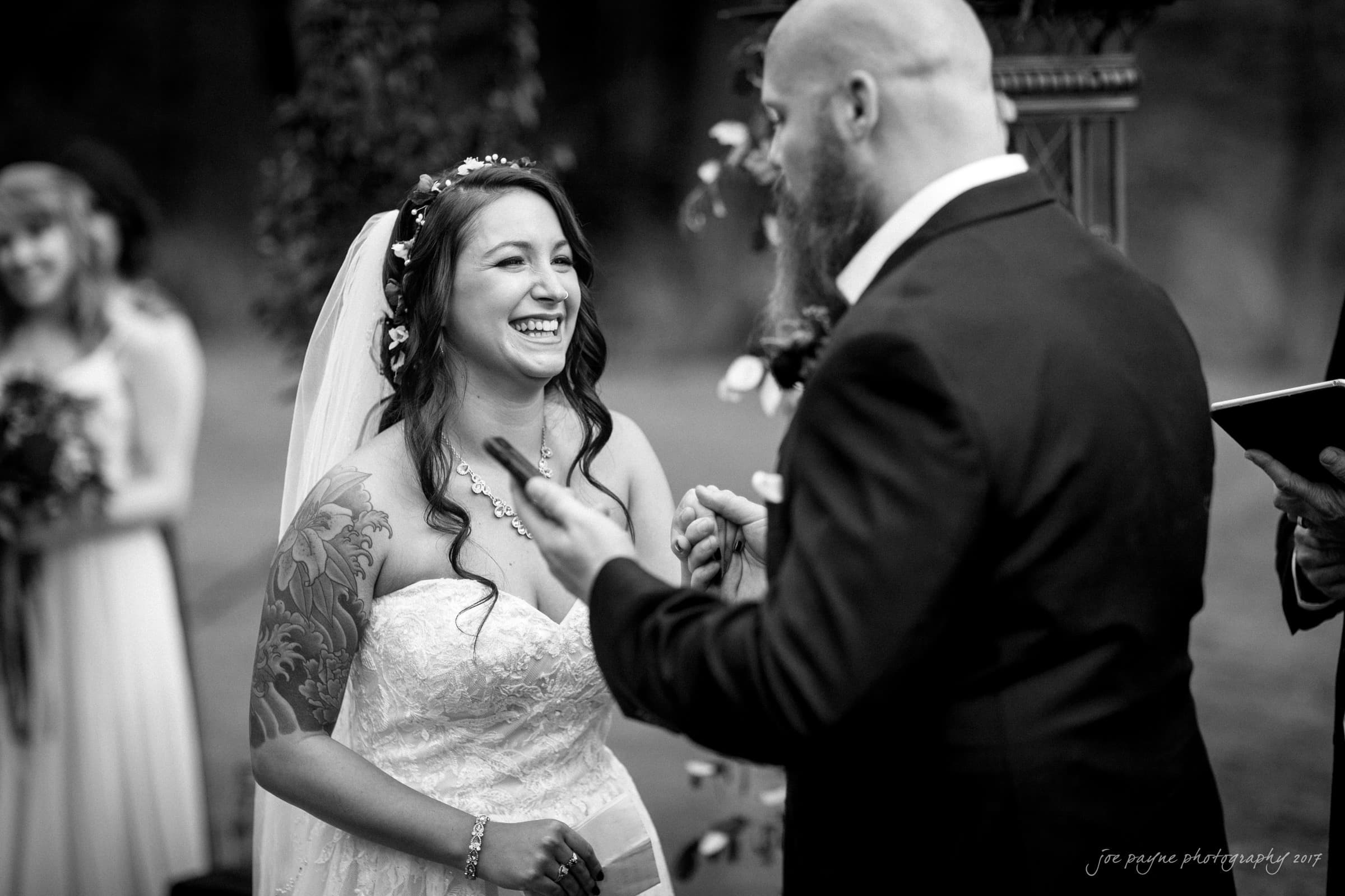 river landing country club wedding - megan & chad A joyful bride in a strapless wedding dress and veil smiles broadly at a bearded groom in a suit during their outdoor wedding ceremony. A bridesmaid holds flowers in the background. Black and white photo capturing a lively moment.
