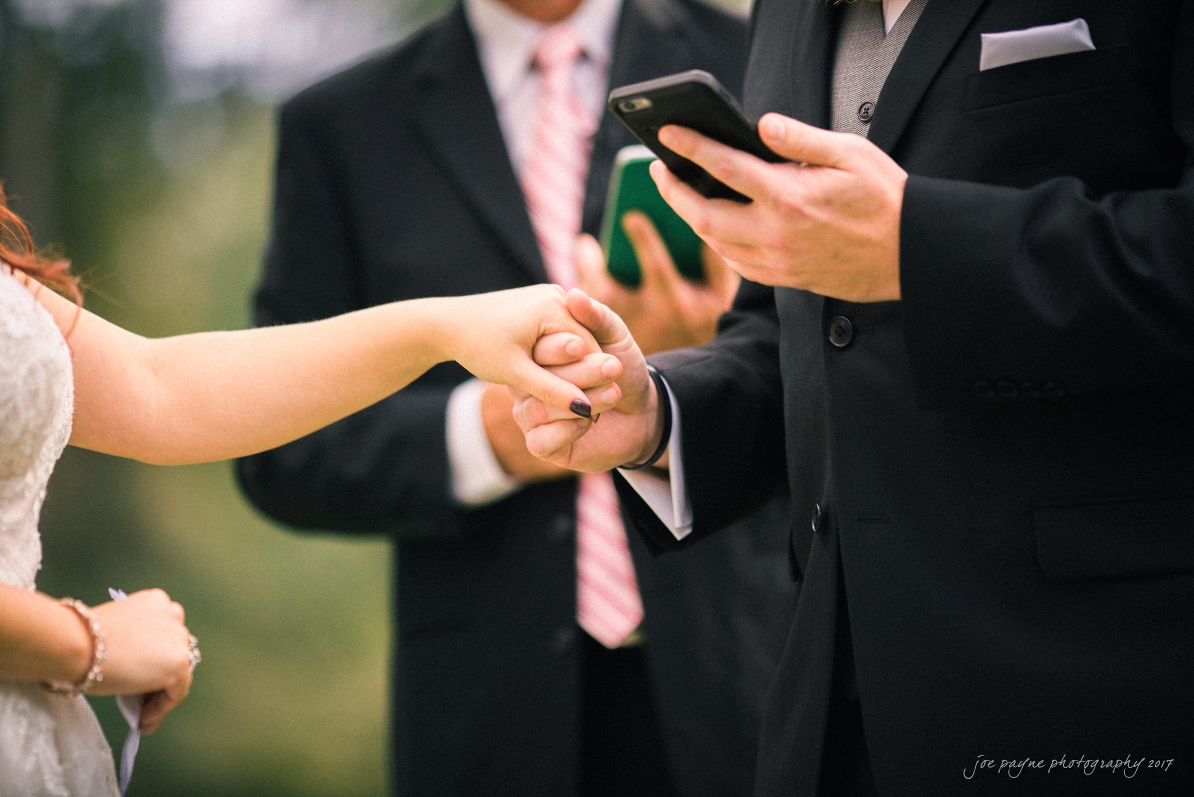 river landing country club wedding - megan & chad A groom, in a black suit, holds a brides hand during a wedding ceremony. The groom reads from a smartphone. The officiant, wearing a suit and tie, stands behind them, slightly out of focus.