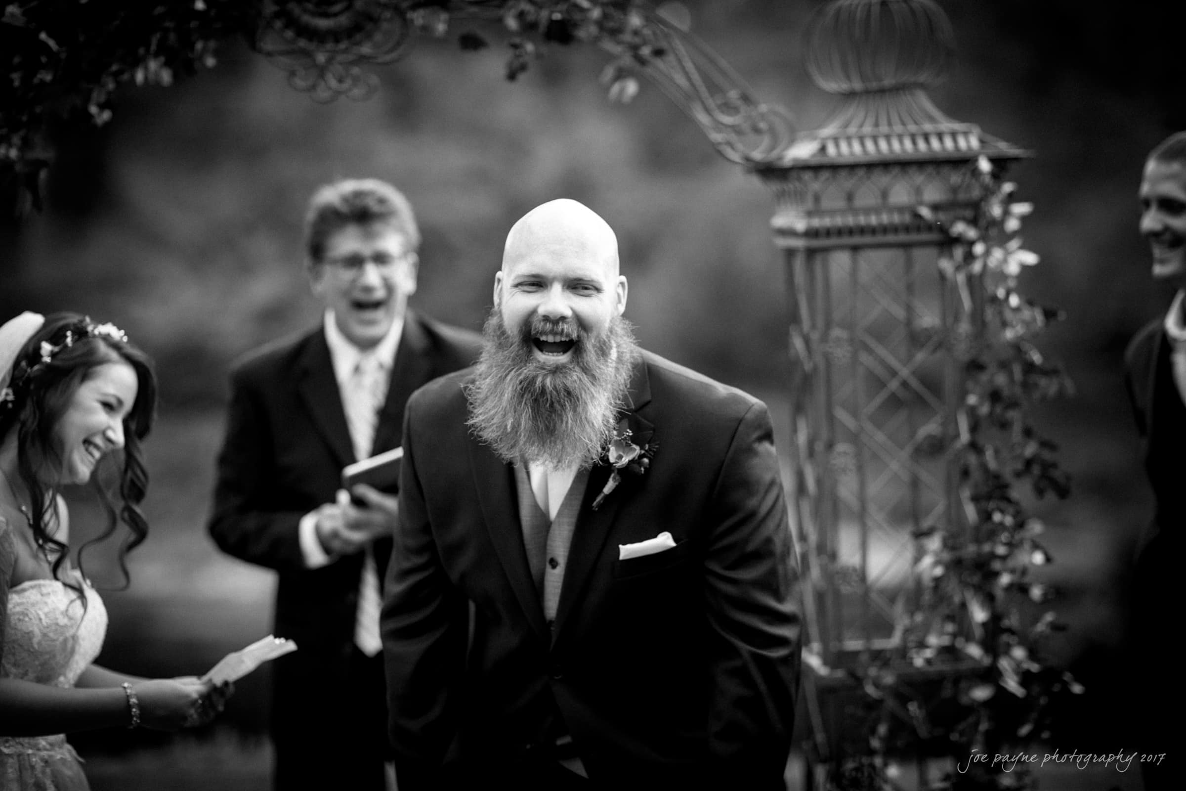 river landing country club wedding - megan & chad A bearded man in a suit laughs during an outdoor wedding ceremony. A woman in a wedding dress and a man holding a book are also laughing. They are standing under an ornate, decorated arch. The image is in black and white.