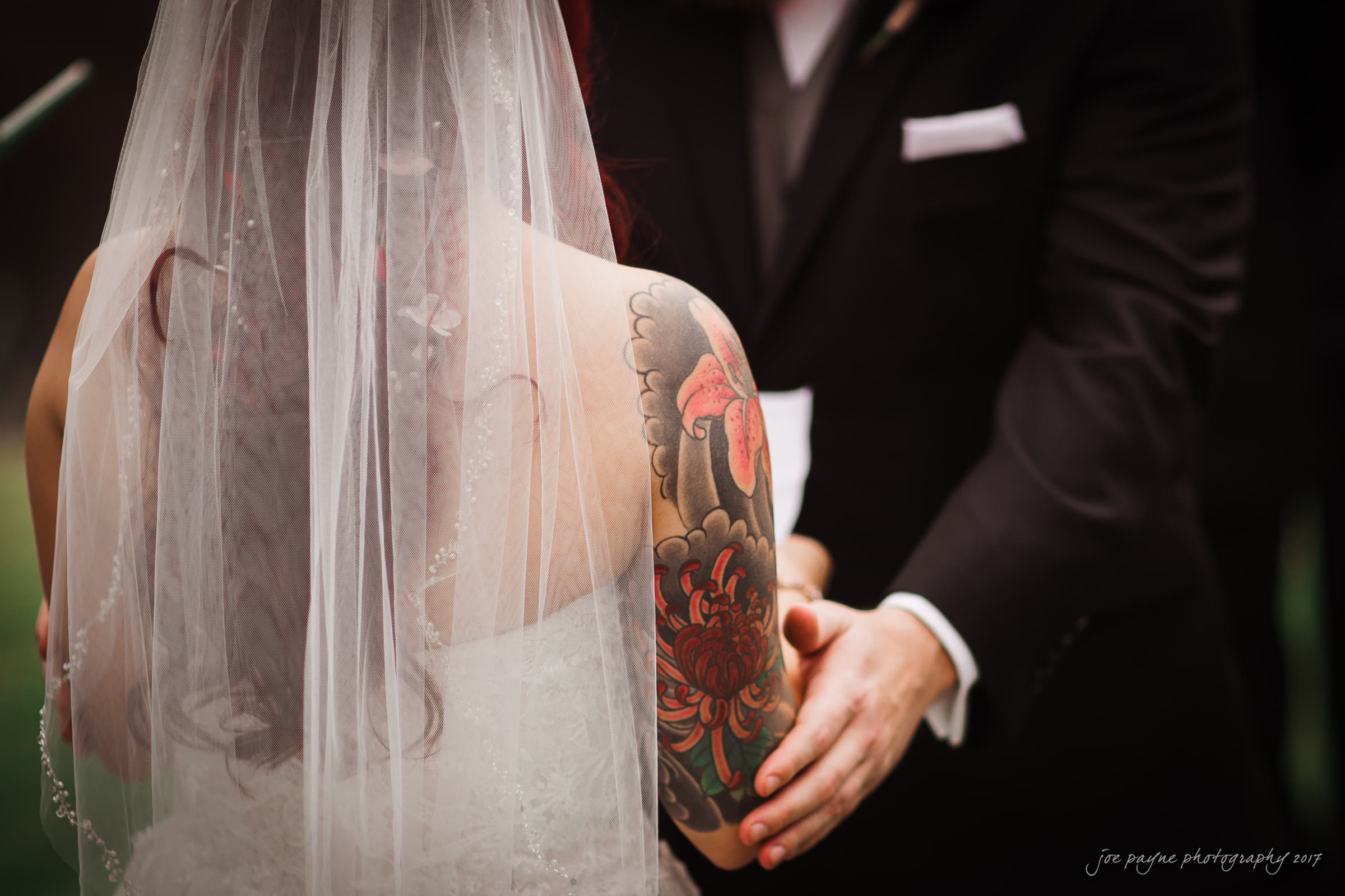 river landing country club wedding - megan & chad A bride and groom hold hands during a wedding ceremony. The brides back is visible, showing a colorful tattoo on her arm. She wears a white veil, and the groom is in a black suit.