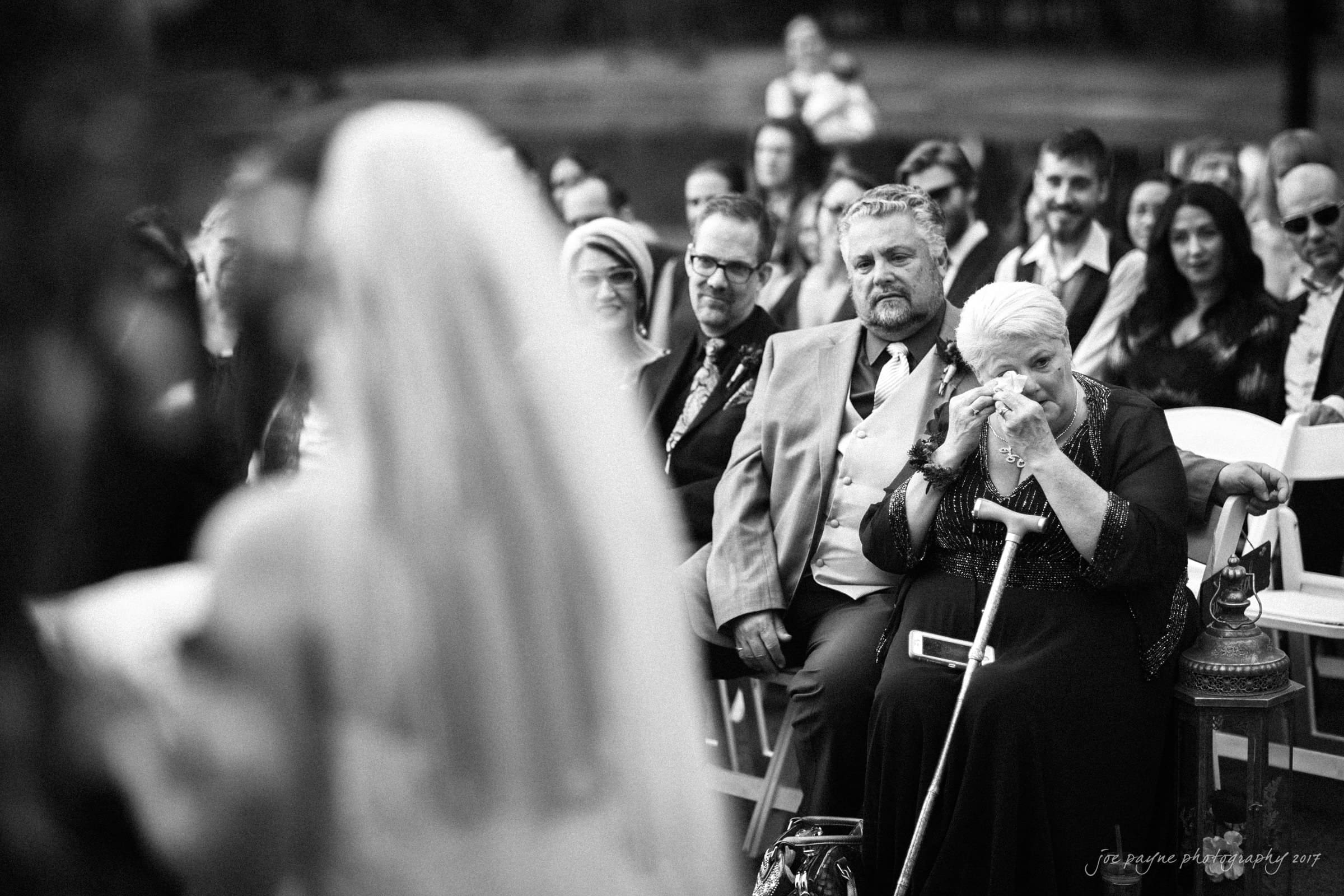 river landing country club wedding - megan & chad A black and white photo shows a seated woman wiping her eyes with a tissue at an outdoor wedding ceremony. Other guests, dressed formally, sit beside her, watching the scene unfold. The blurred figure of the bride is in the foreground.