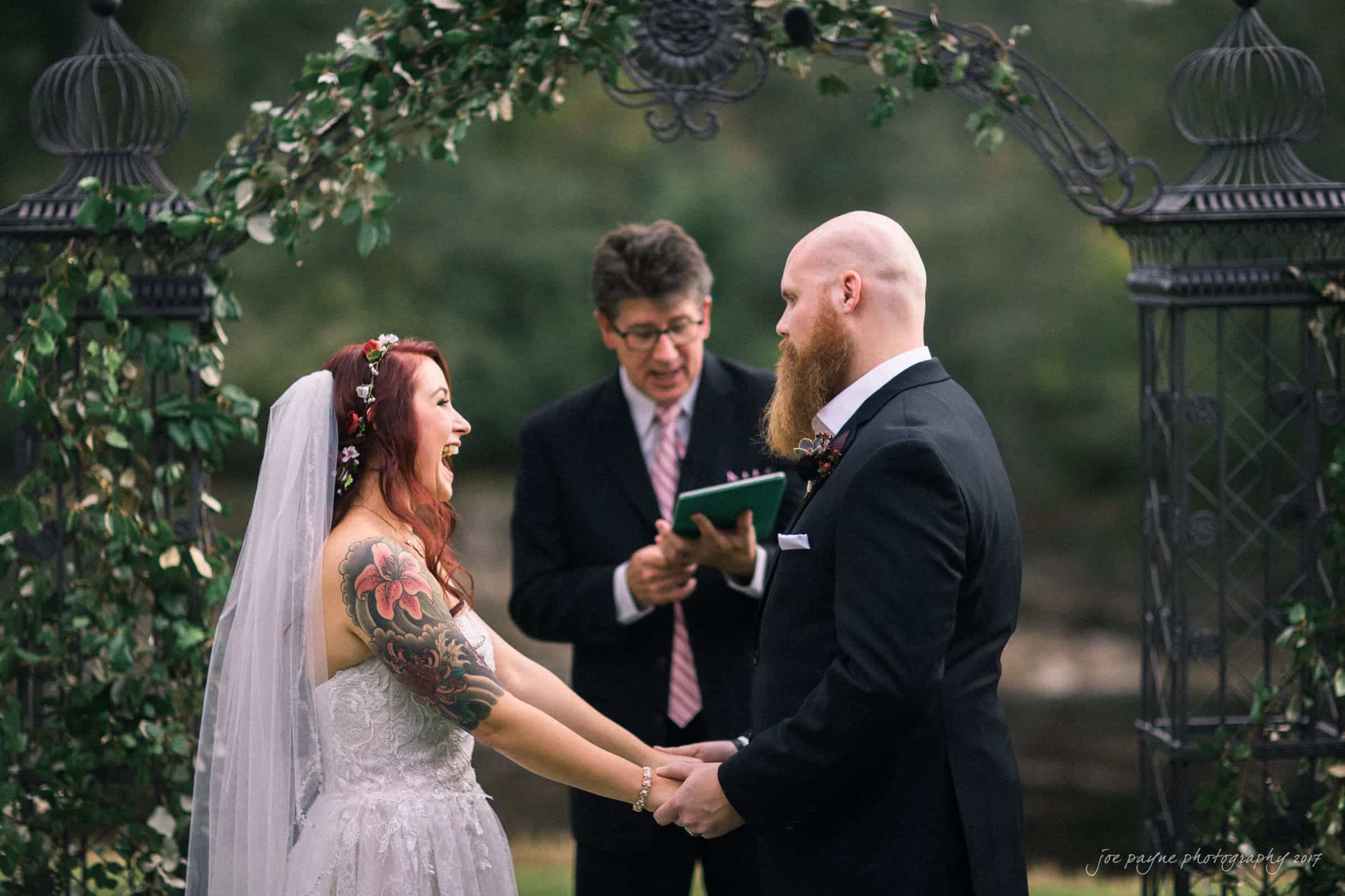 river landing country club wedding - megan & chad A bride in a strapless white gown and floral crown holds hands with a groom in a black suit. They stand under an arch decorated with greenery. A person in formal attire officiates the wedding, holding a book. The couple smiles at each other.