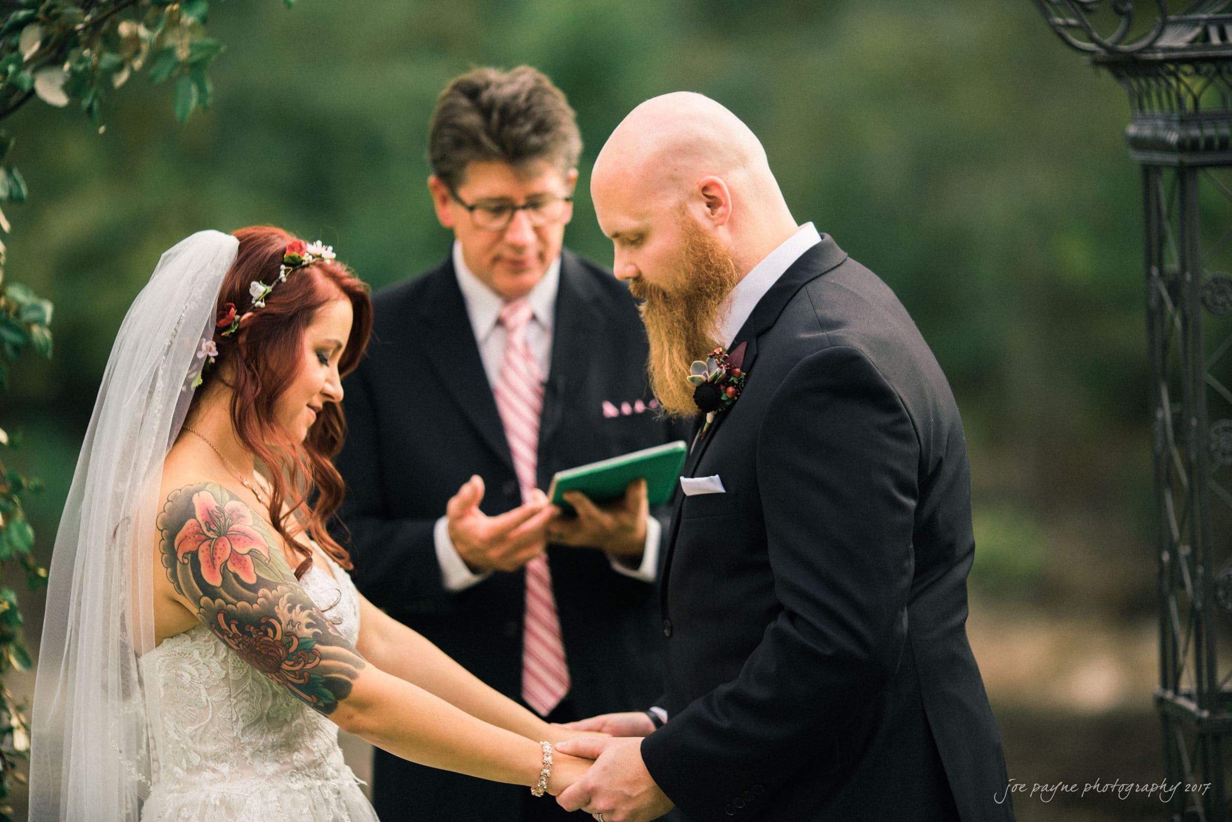 river landing country club wedding - megan & chad A couple stands facing each other, holding hands in a wedding ceremony. The bride wears a floral headpiece and a white dress with tattoos on her arm. The groom wears a black suit. An officiant stands behind them holding a book. Lush greenery surrounds them.