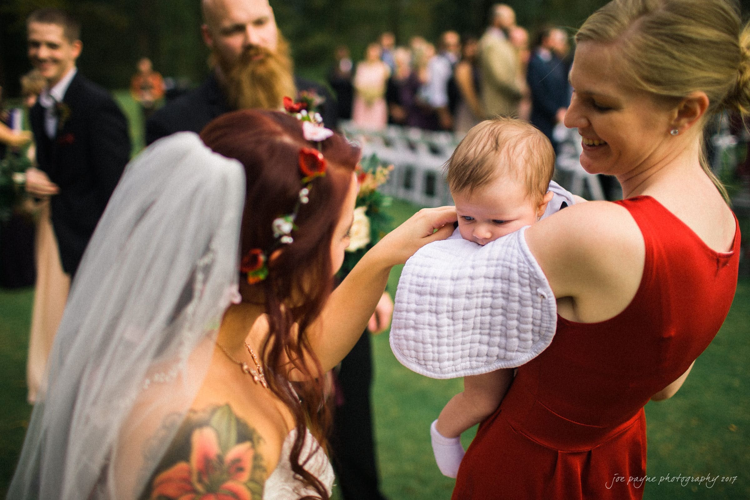 river landing country club wedding - megan & chad A bride wearing a floral crown smiles at a baby held by a woman in a red dress. The scene is outdoors with guests and greenery in the background. The baby is wearing a white bib and looking curiously at the bride.