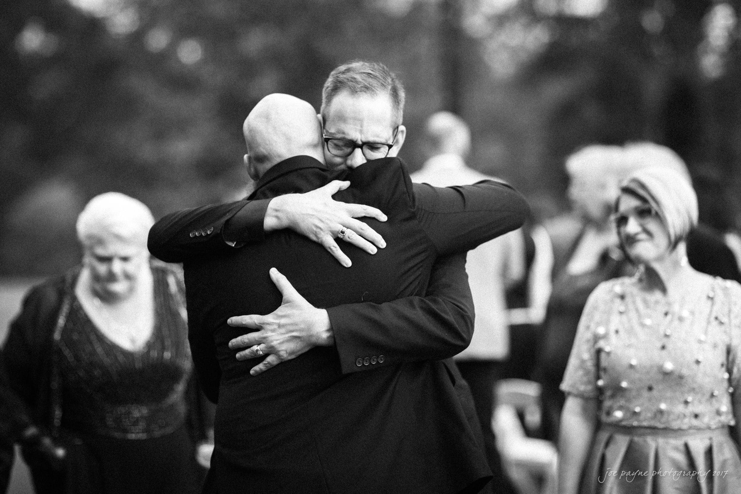 river landing country club wedding - megan & chad A black and white photo of two men embracing warmly, with one facing the camera. A group of people stands in the background, including a woman on the left and another on the right, all appearing to be at an outdoor gathering.