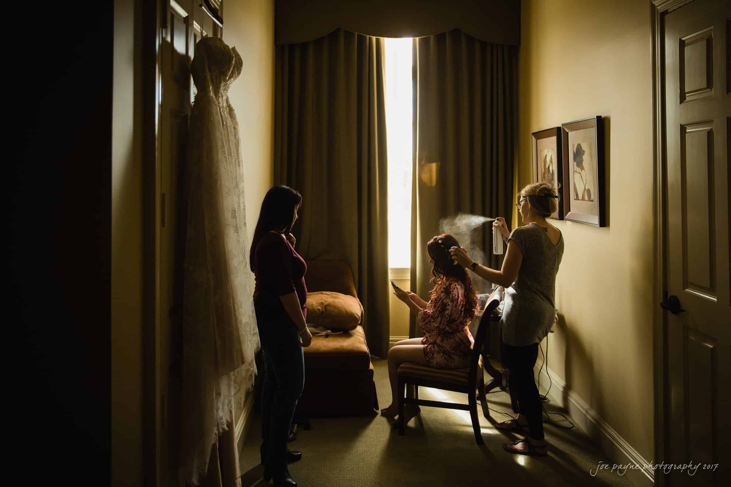 river landing country club wedding - megan & chad A bride in a robe sits in a chair getting her hair styled by a stylist using a curling iron. Another woman stands nearby. The room is dimly lit, with a wedding dress hanging on the wall and light streaming through the window.