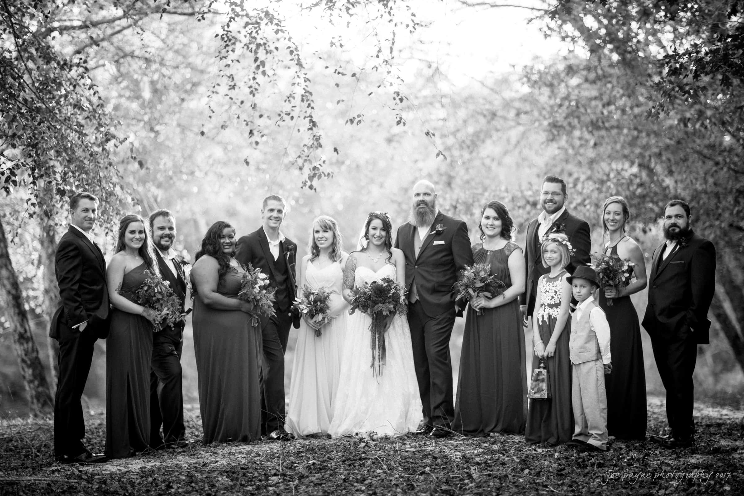 river landing country club wedding - megan & chad A black and white photo of a wedding party in a wooded area. The group includes the bride, groom, bridesmaids, groomsmen, and children. They are dressed in formal attire, standing in a line with trees in the background.
