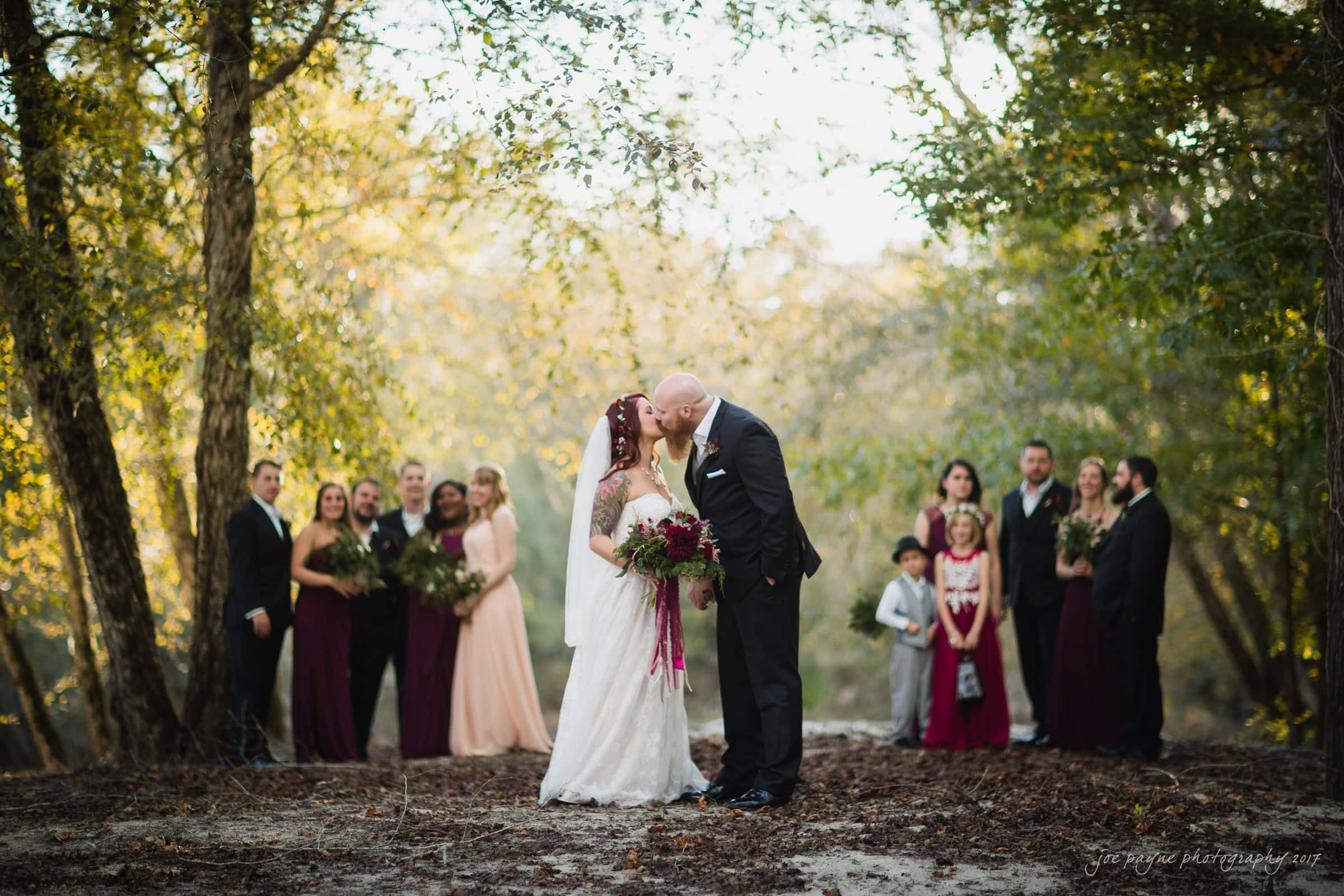 river landing country club wedding - megan & chad A bride and groom share a kiss in a forest clearing, surrounded by their wedding party. Bridesmaids in pink dresses and groomsmen in dark suits stand on either side, with trees and soft sunlight in the background.