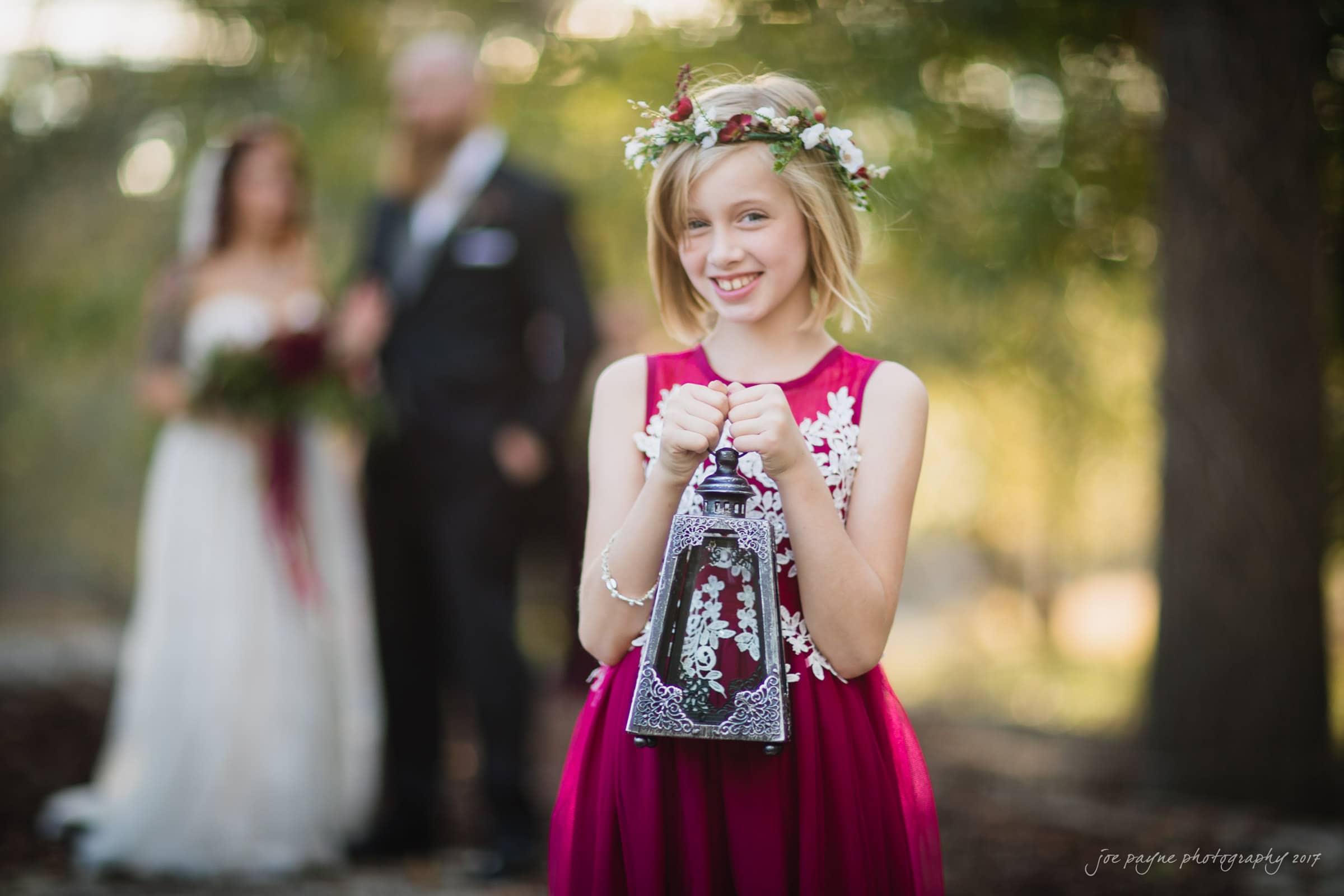 river landing country club wedding - megan & chad A young girl in a bright pink dress and floral headband smiles while holding a decorative lantern. In the blurred background, a couple stands together, with the woman in a wedding dress and the man in a suit, outdoors with trees around them.