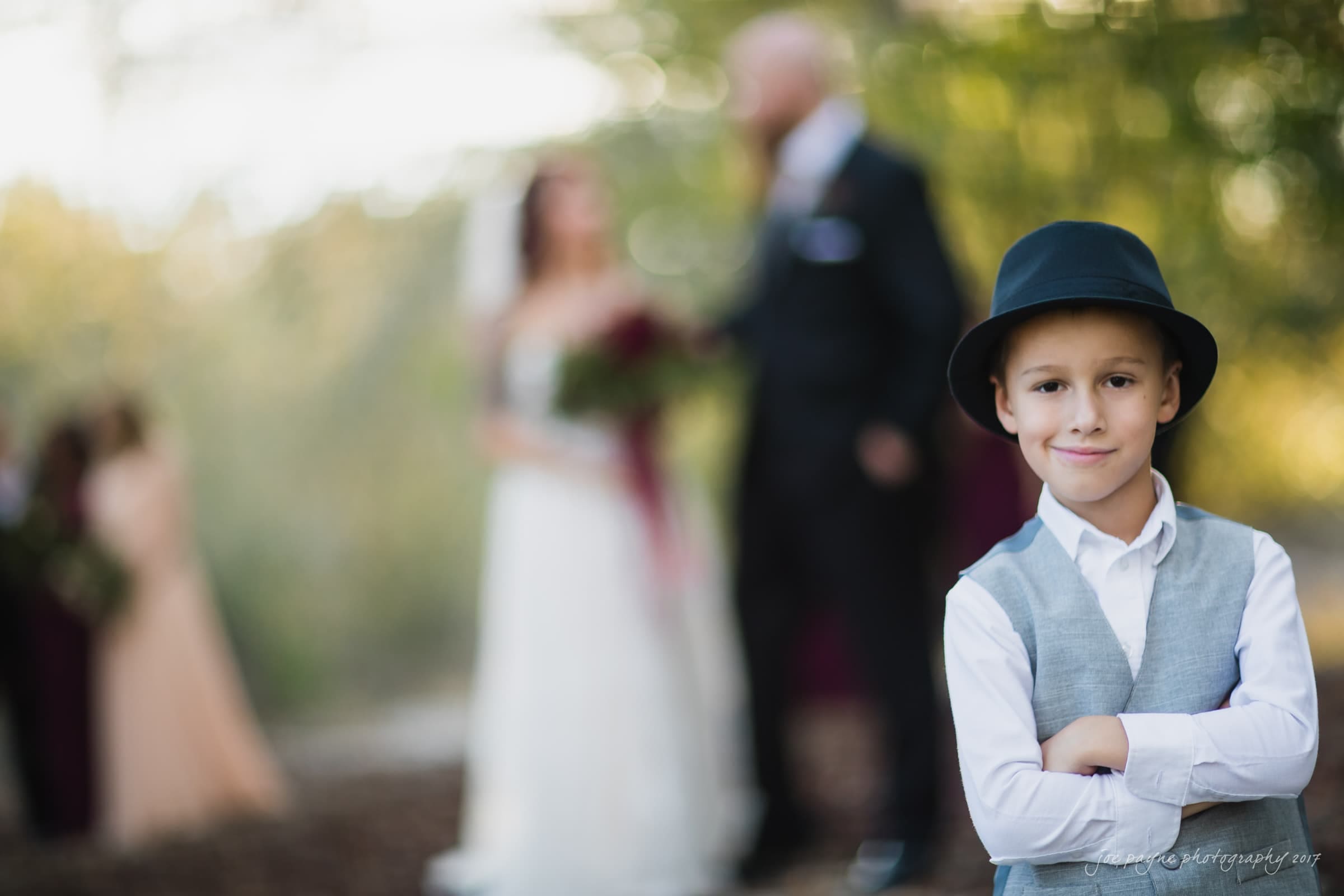 river landing country club wedding - megan & chad A young boy in a grey vest and black hat stands with arms crossed, smiling at the camera. In the blurred background, a bride and groom stand together, with bridesmaids visible on the side amidst a natural, wooded setting.