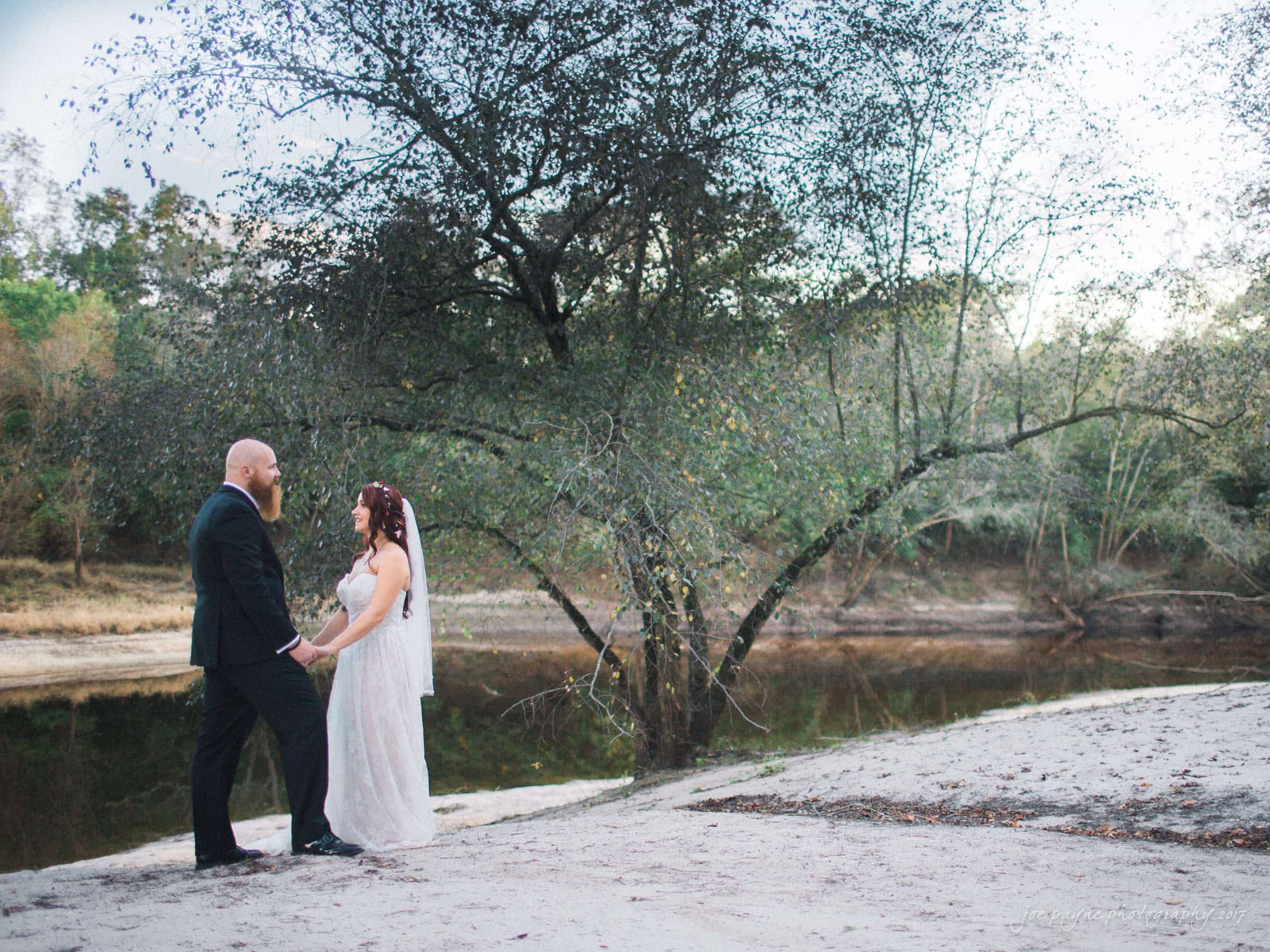 river landing country club wedding - megan & chad A bride and groom stand facing each other by a tranquil riverbank. The groom is wearing a black suit, and the bride is in a flowing white dress. They are surrounded by trees with sparse leaves under a cloudy sky. The scene is peaceful and natural.