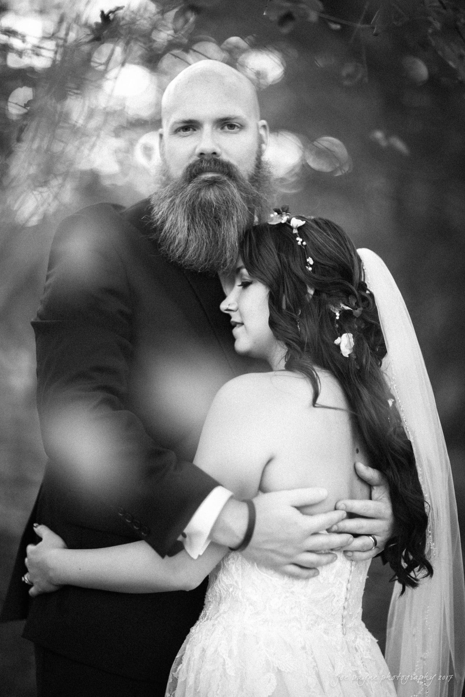 river landing country club wedding - megan & chad A black and white photo of a groom with a beard and a bride wearing a floral headpiece and veil. They are embracing, with her eyes closed and his looking at the camera, surrounded by a blurred, nature-filled background.
