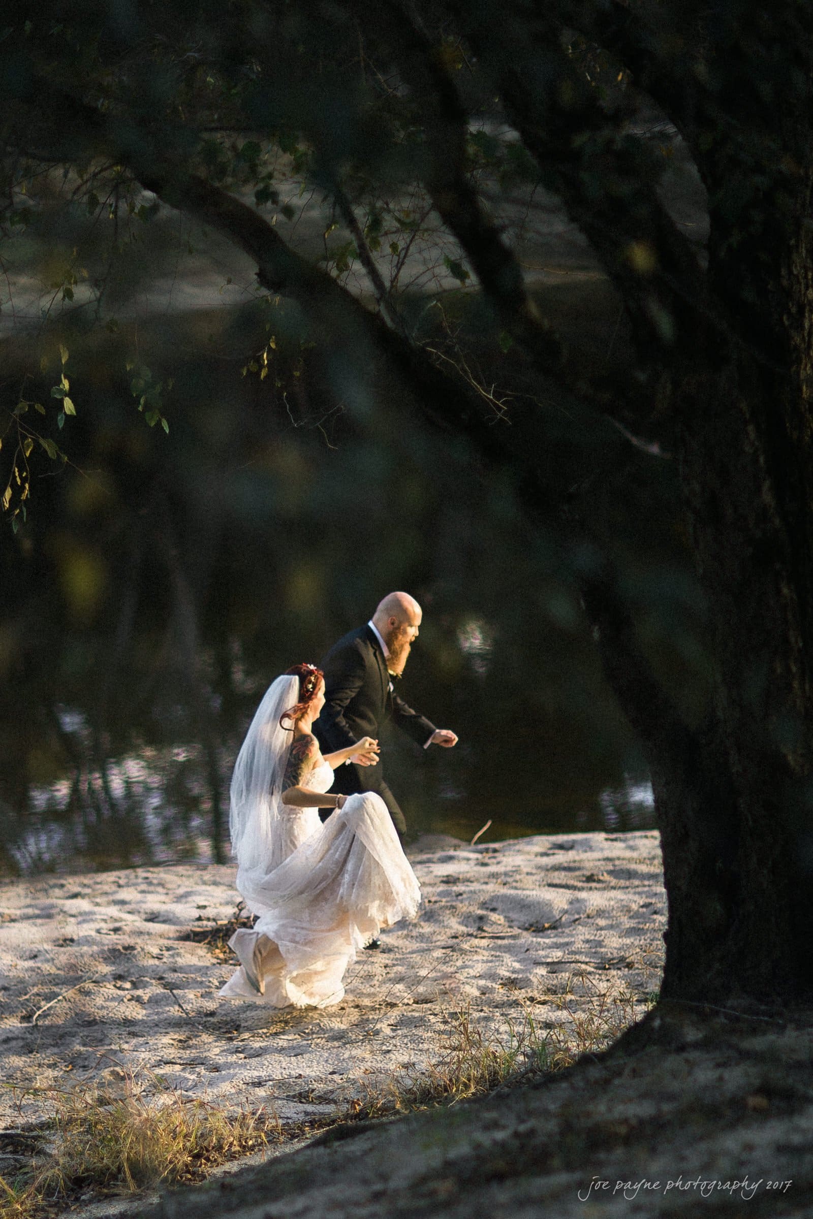 river landing country club wedding - megan & chad A bride in a white dress and veil walks alongside a man in a dark suit. They are on a sandy path by the water, framed by tree branches, with sunlight filtering through.